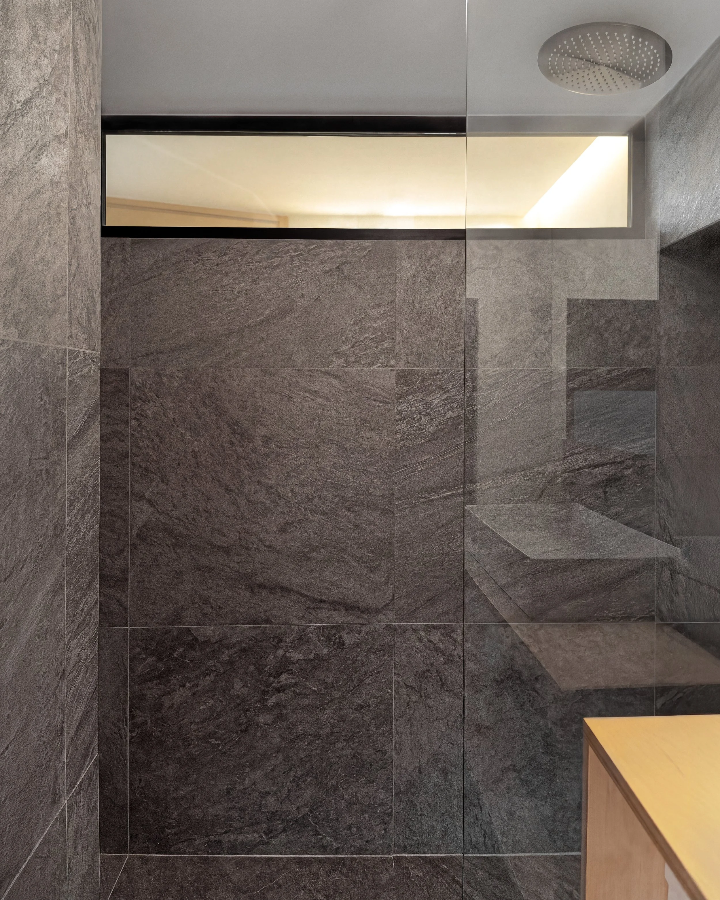 The image shows a modern, sleek shower area with dark stone tiles on the walls and floor. Above, there is a ceiling with a built-in rainfall showerhead. On the right side, a small wooden table is visible.