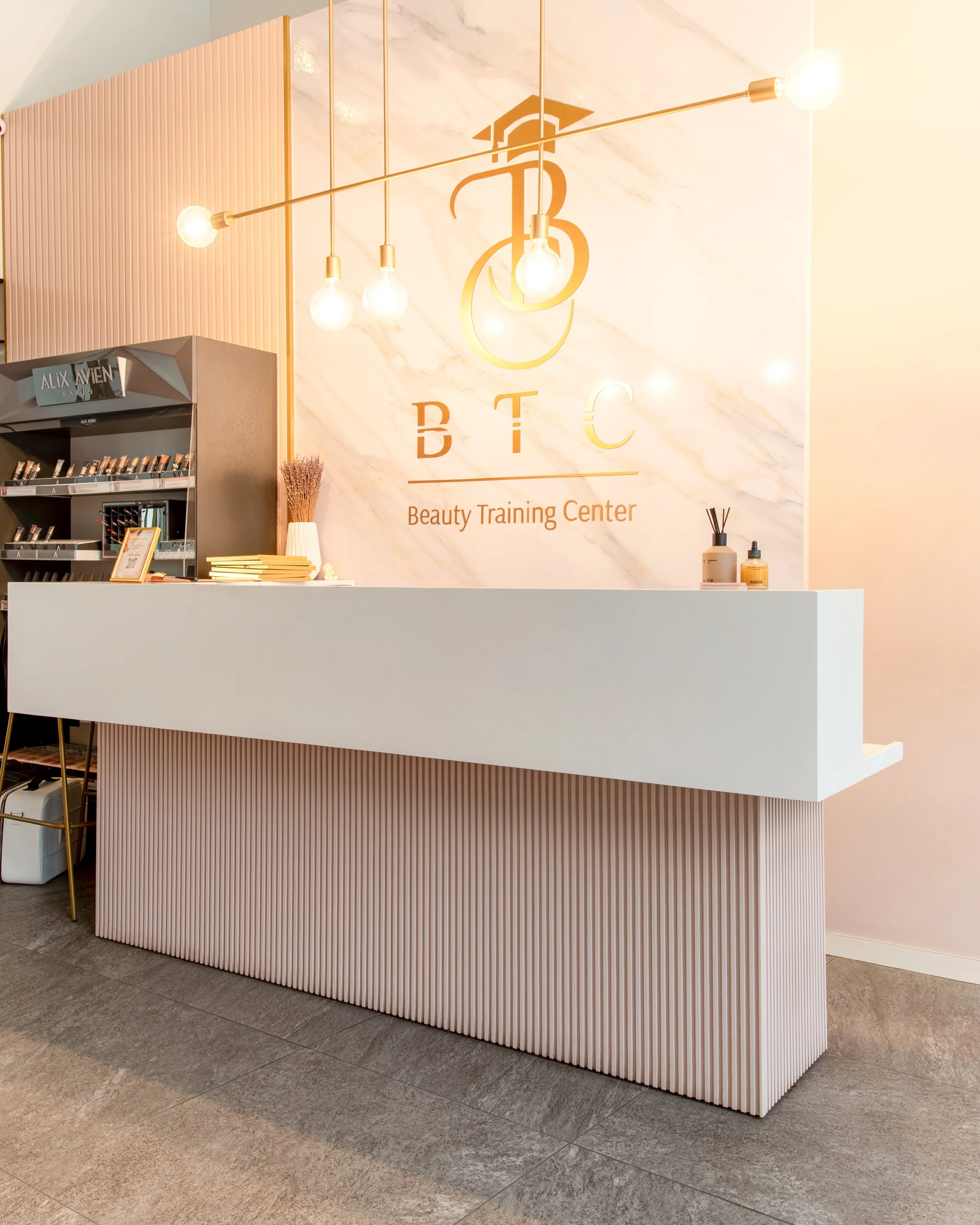 Reception area of beauty training center with a white marble wall featuring a logo, a white counter with makeup products, small decorative plants, and a pink ribbed panel on the side, illuminated by a gold hanging light fixture with four bulbs.