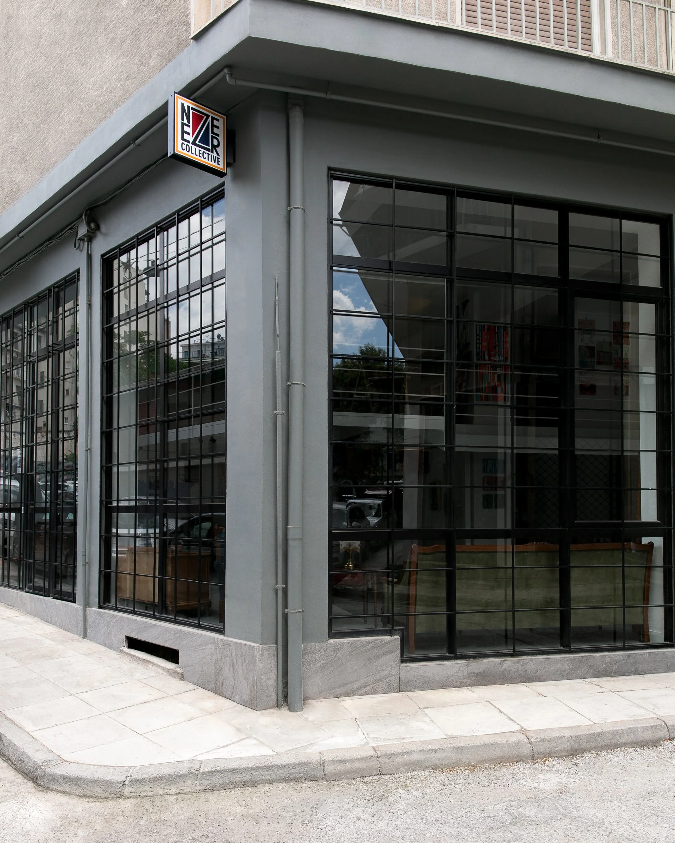 The image shows the corner of a modern building with large glass windows and a sign that reads 'NEZ Collective' on the upper part of the corner. The building has a gray exterior and is located at the intersection with a paved sidewalk and street.
