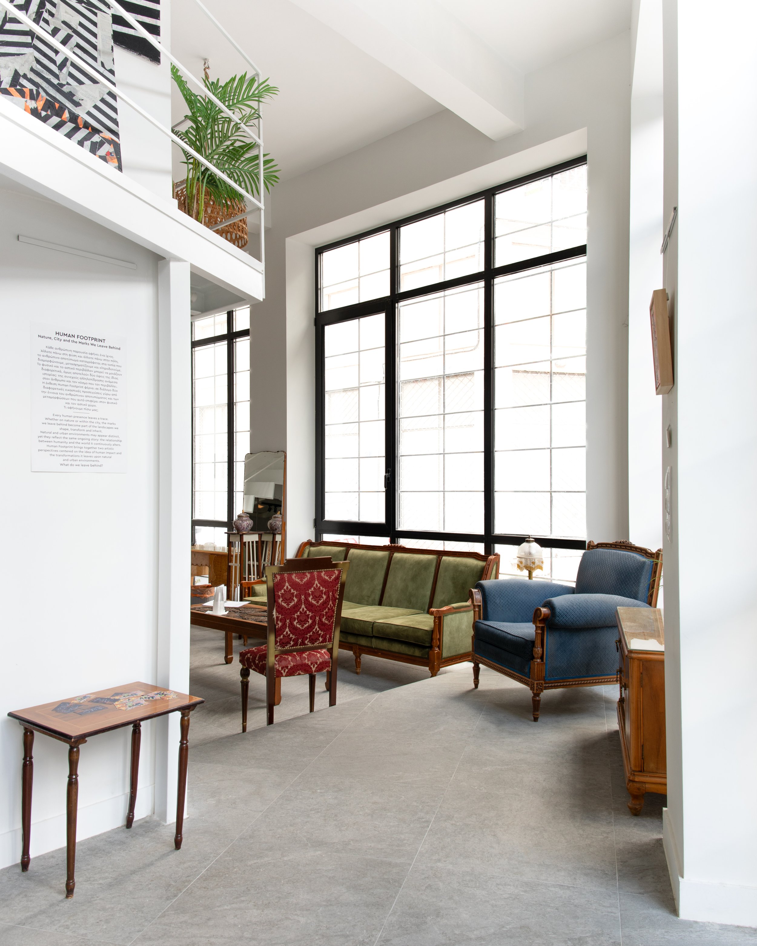 Interior of a modern building with large windows, classic furniture including a green velvet sofa, red patterned armchair, blue armchair, and a wooden coffee table. A white wall has a sign titled 'Human Footprint' with text, and there are decorative 
