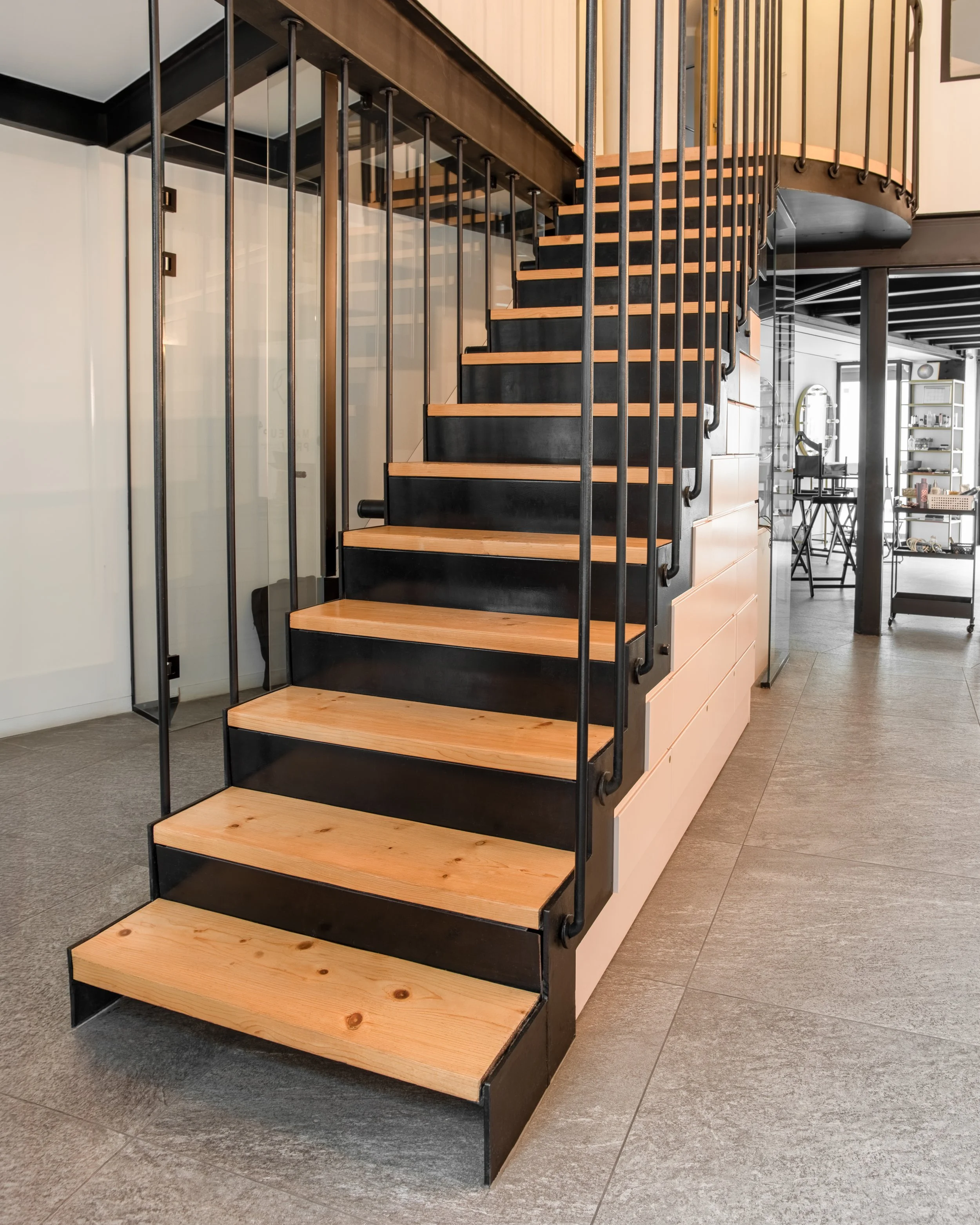 A modern spiral staircase with black treads and wooden edges, glass and metal railing, in a contemporary interior space.