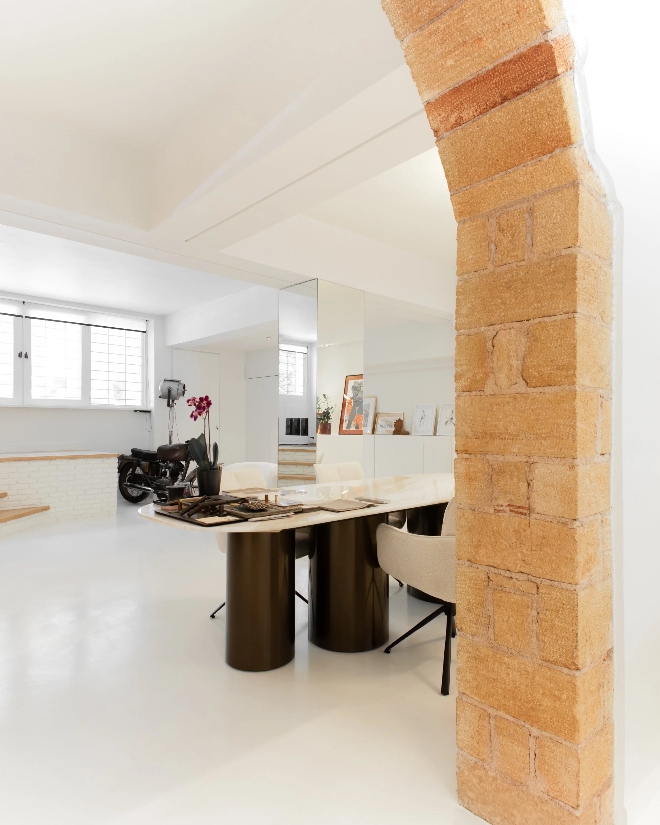 Modern dining room with white walls, large windows, black cylindrical dining table, beige upholstered chairs, stacked books, decorative items, and brick column.