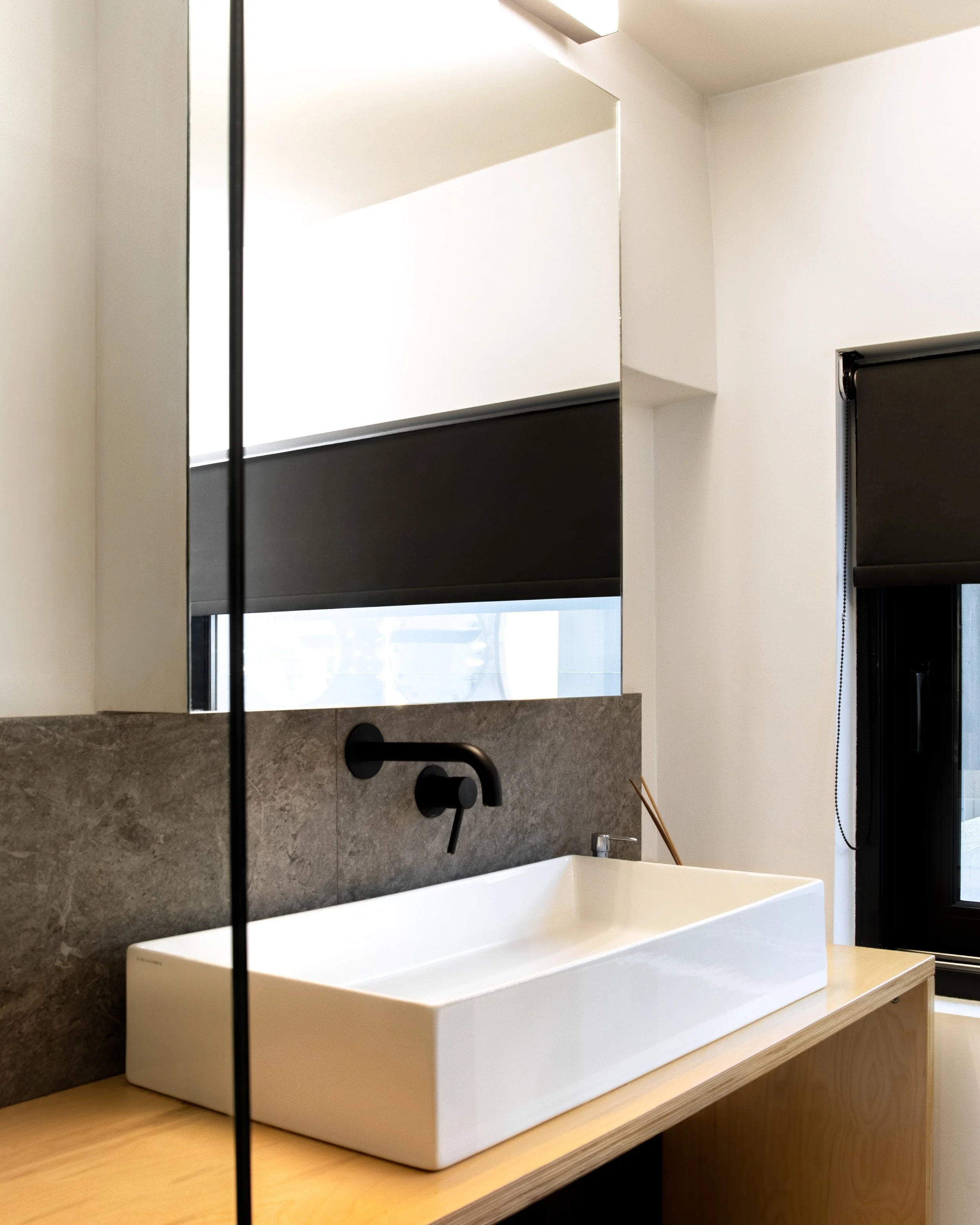 Modern bathroom with a white rectangular sink on a wooden countertop, black wall-mounted faucet, grey stone wall, large mirror, and a window with black roller shade.