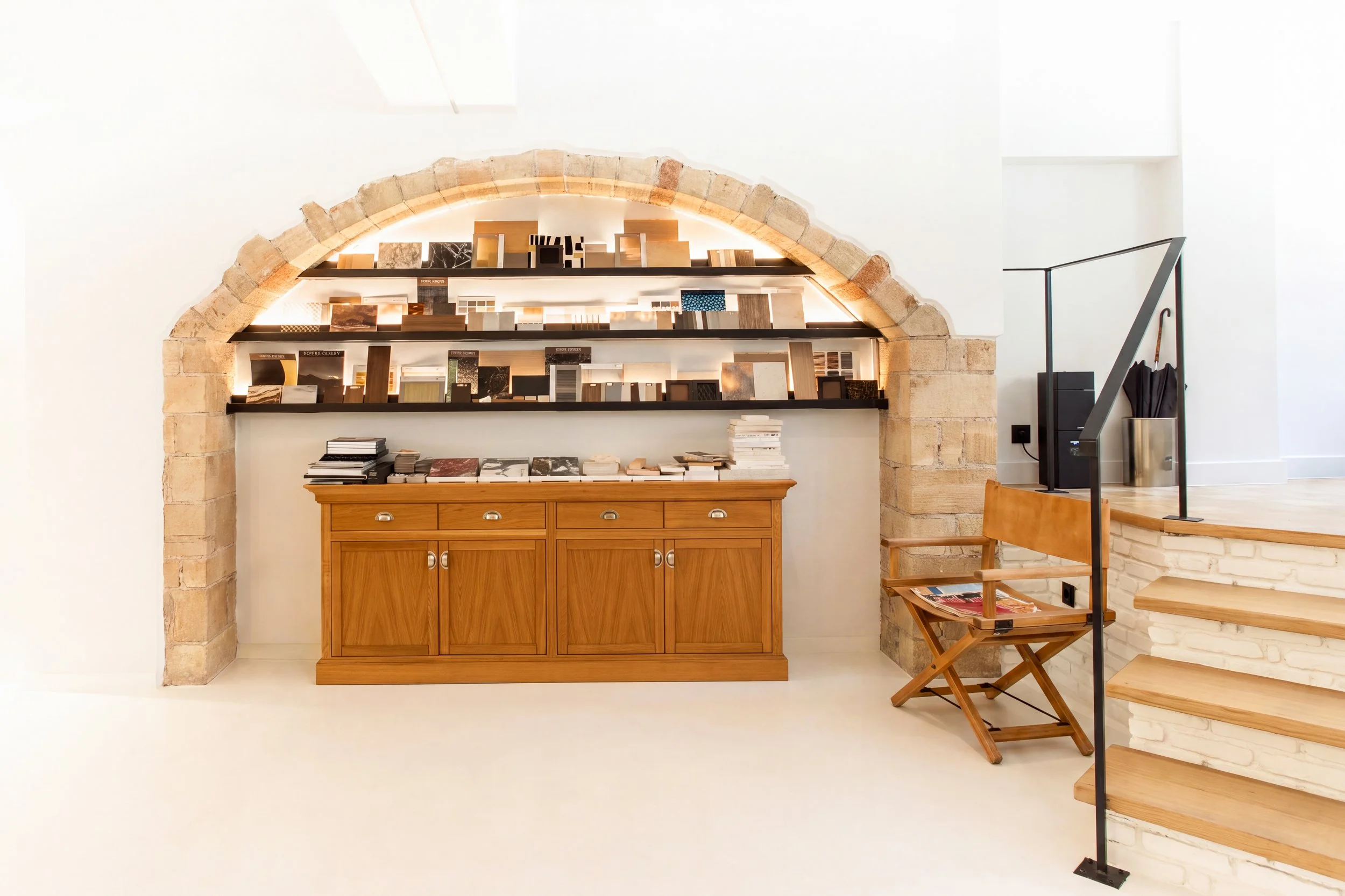 Interior view of a modern room with a stone archway, wooden cabinet, and black shelves displaying various sample materials, with a staircase and chair in the background.