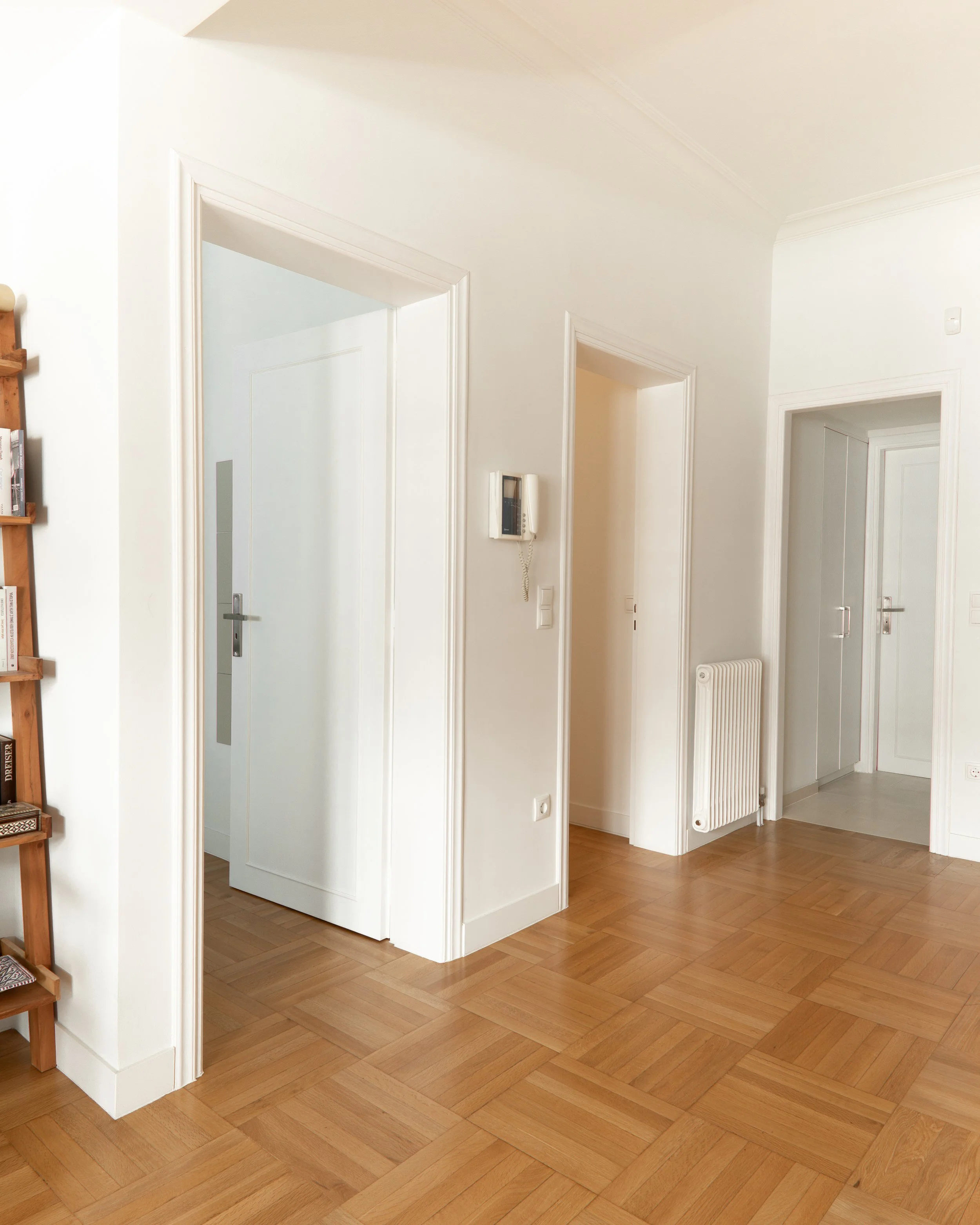 Interior of a house with wooden flooring, white walls, and multiple doorways. There is a wall-mounted telephone on the wall, and a radiator near the doorways. Part of a wooden bookshelf or ladder is visible on the left side.
