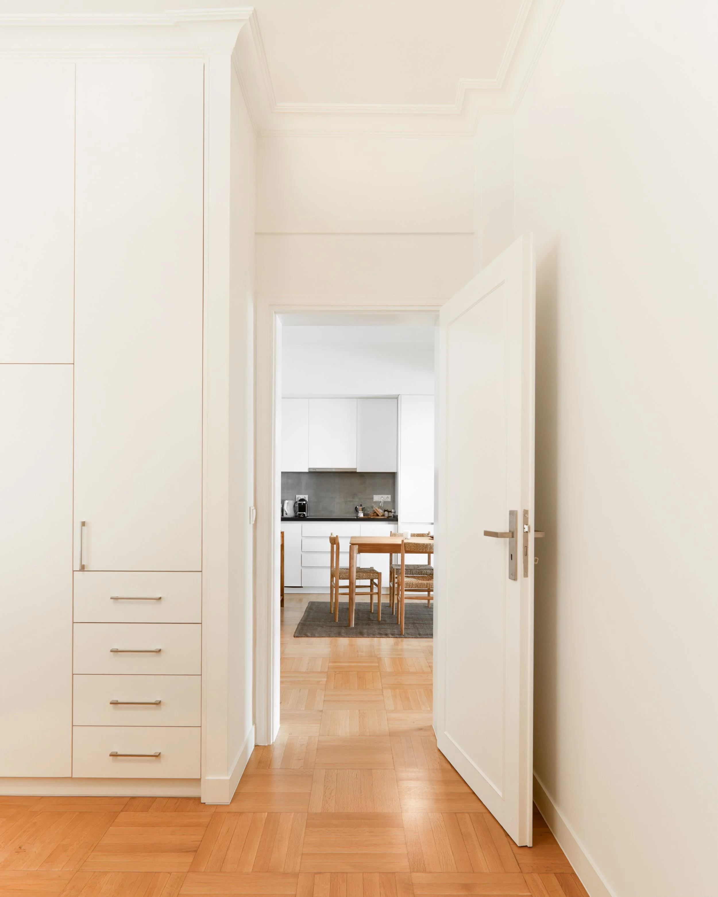 A hallway leading to a kitchen with a dining table and chairs, viewed through a doorway, with light wood flooring and white walls.