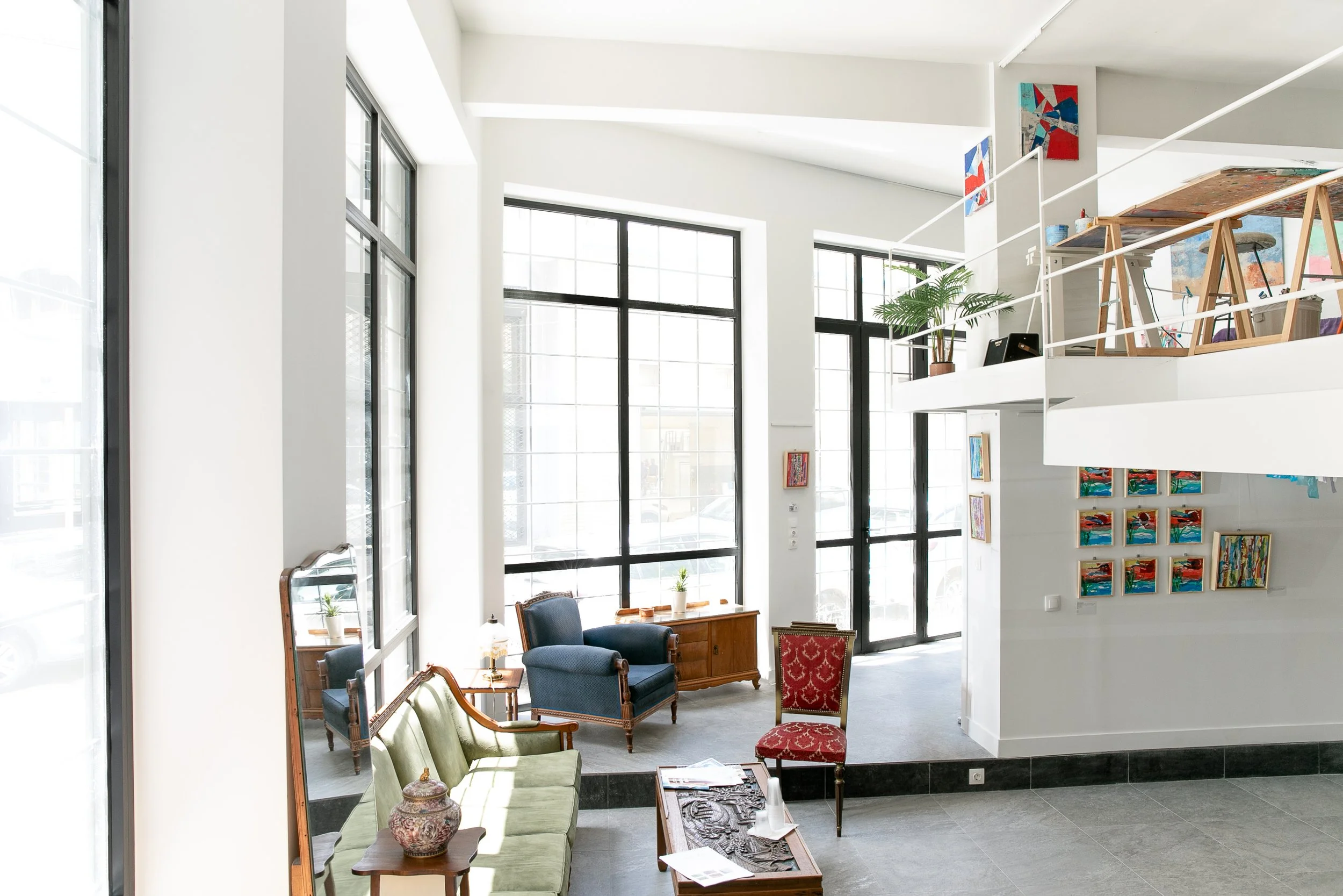 A bright, open indoor space resembling an art gallery or modern living area with large windows, colorful artwork on the walls, and vintage-style furniture including chairs and a sofa.