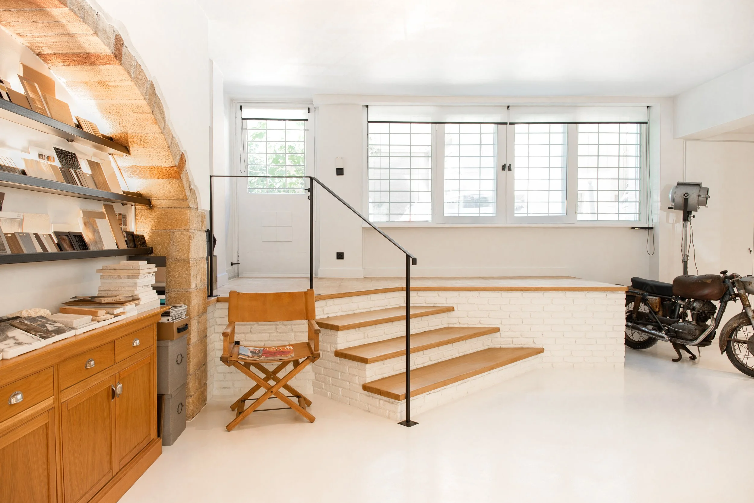 Interior of a bright room with large windows, a staircase with wooden steps, a wooden chair with magazines, a motorcycle, and a wall-mounted spotlight.