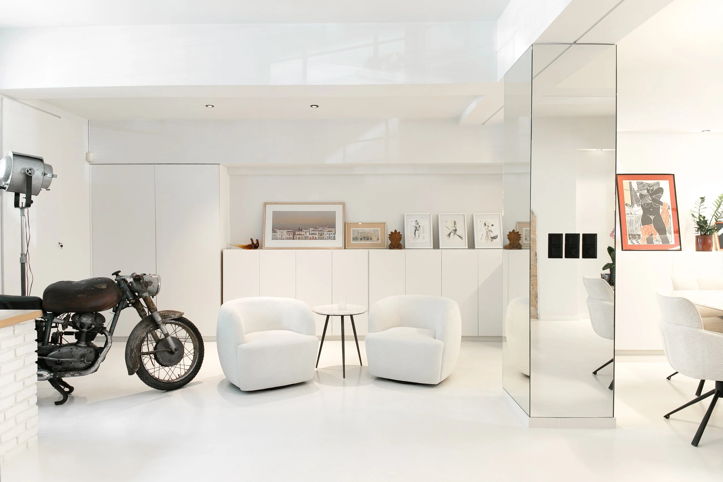 Bright, modern living room with white walls and furnishings, artwork on the wall, a vintage motorcycle, and a studio light