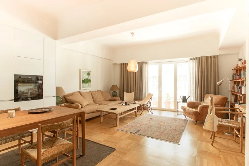 Bright living room with beige sofa, armchair, coffee table, rug, dining table with chairs, bookshelf, and large windows with beige curtains.