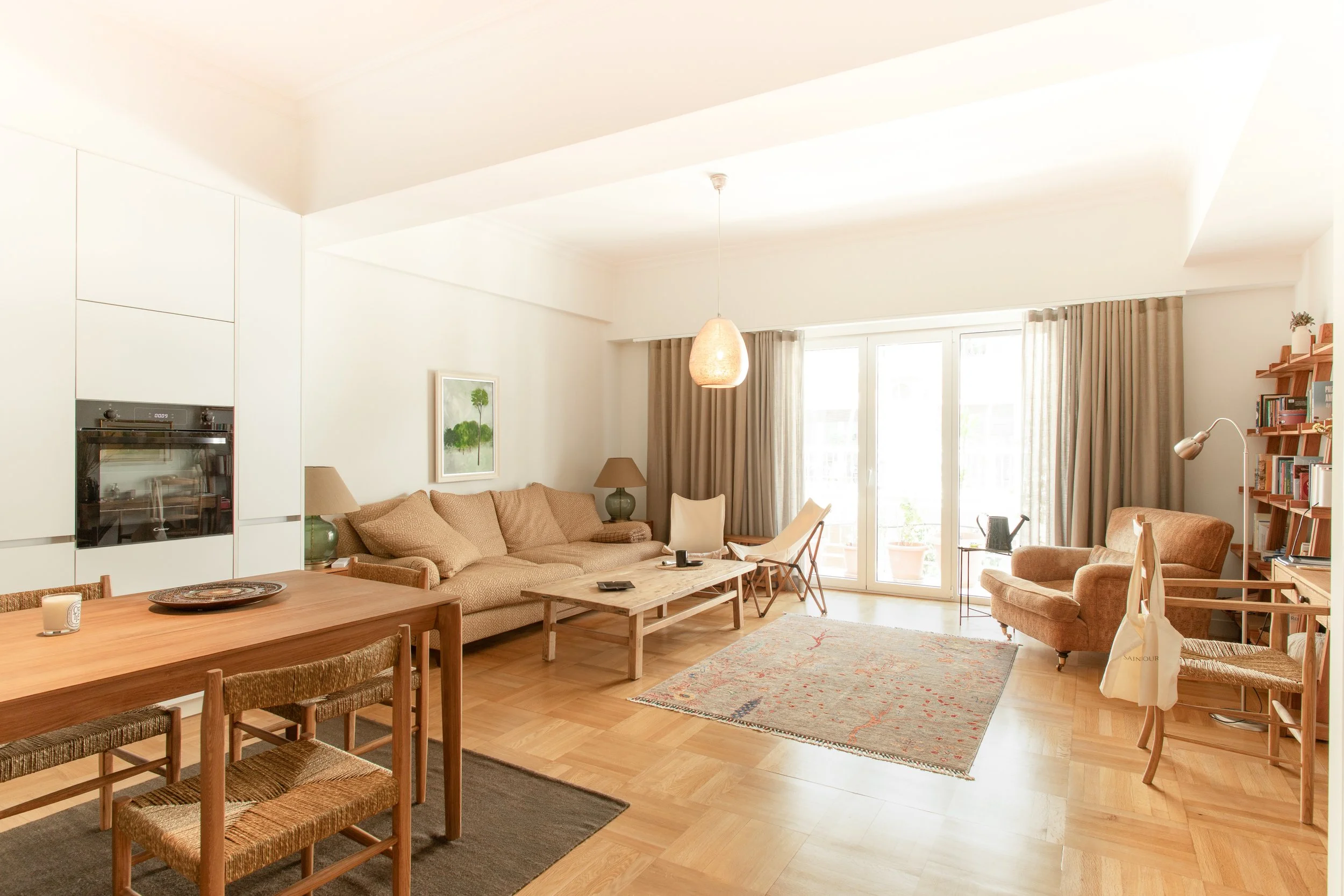 Bright living room with beige couches, wooden dining table with chairs, bookshelf, and large windows with beige curtains.