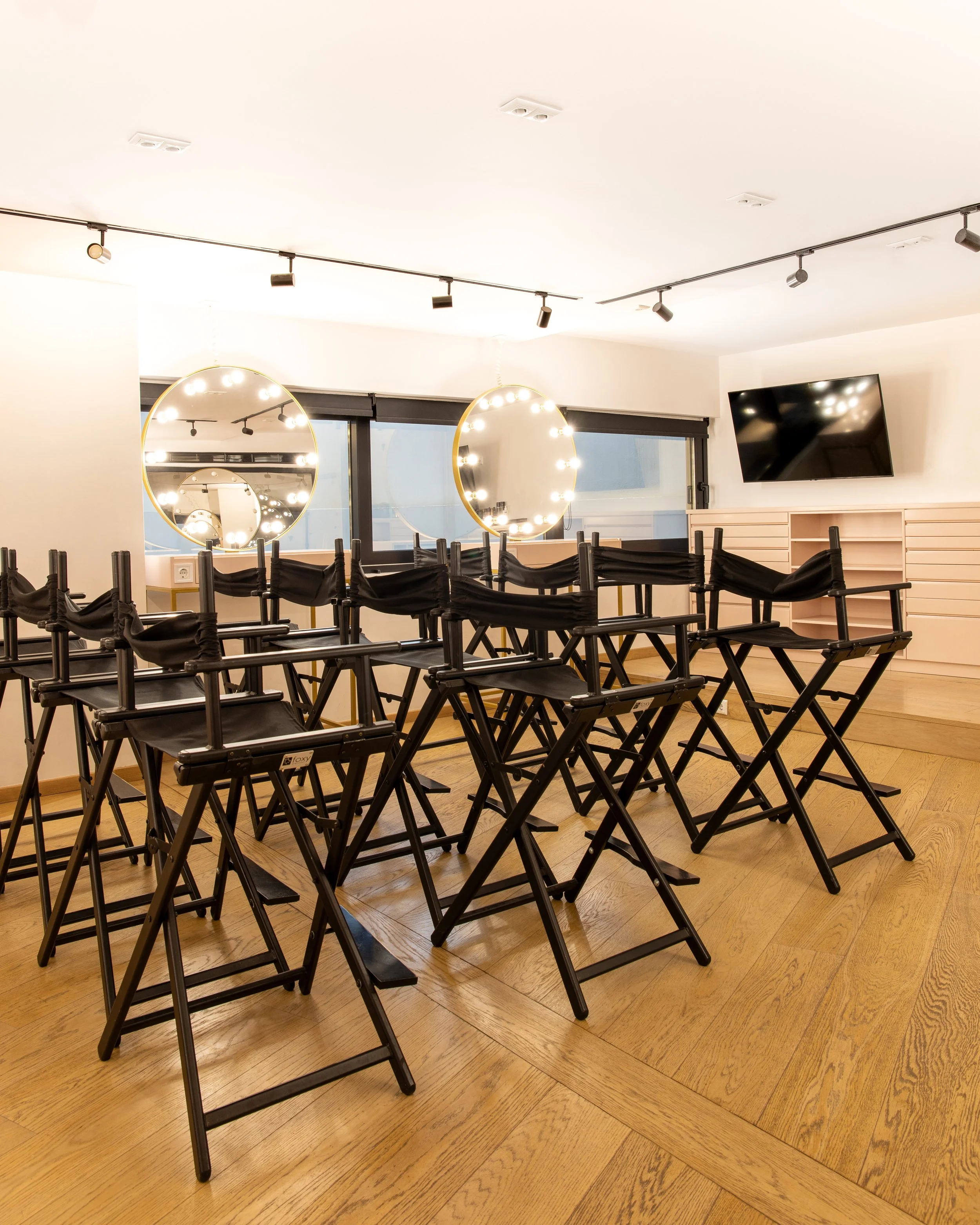 Empty chairs arranged in rows in a room, with mirrors reflecting ceiling lights and a TV mounted on the wall.
