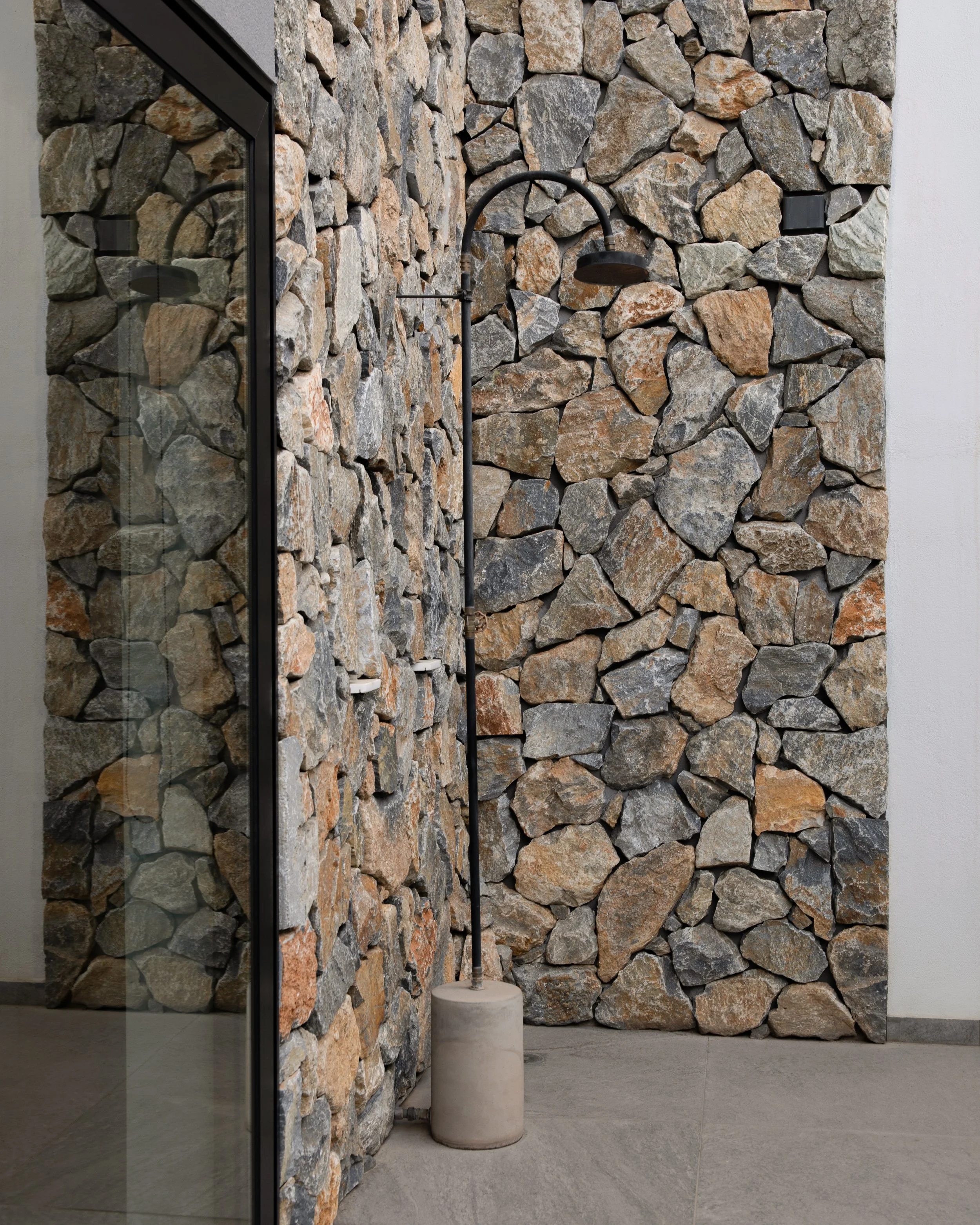 Stone wall with a black outdoor shower fixture and a circular concrete base, adjacent to a glass wall and light gray tiled flooring.