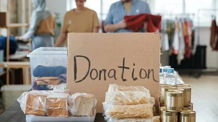 Table with donation items including canned goods, bread, and bottled water, with a cardboard box labeled 'Donation'; background shows people shopping in a thrift store.