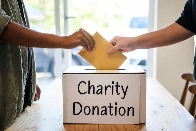 Two people are exchanging an envelope into a donation box labeled 'Charity Donation' on a wooden table.