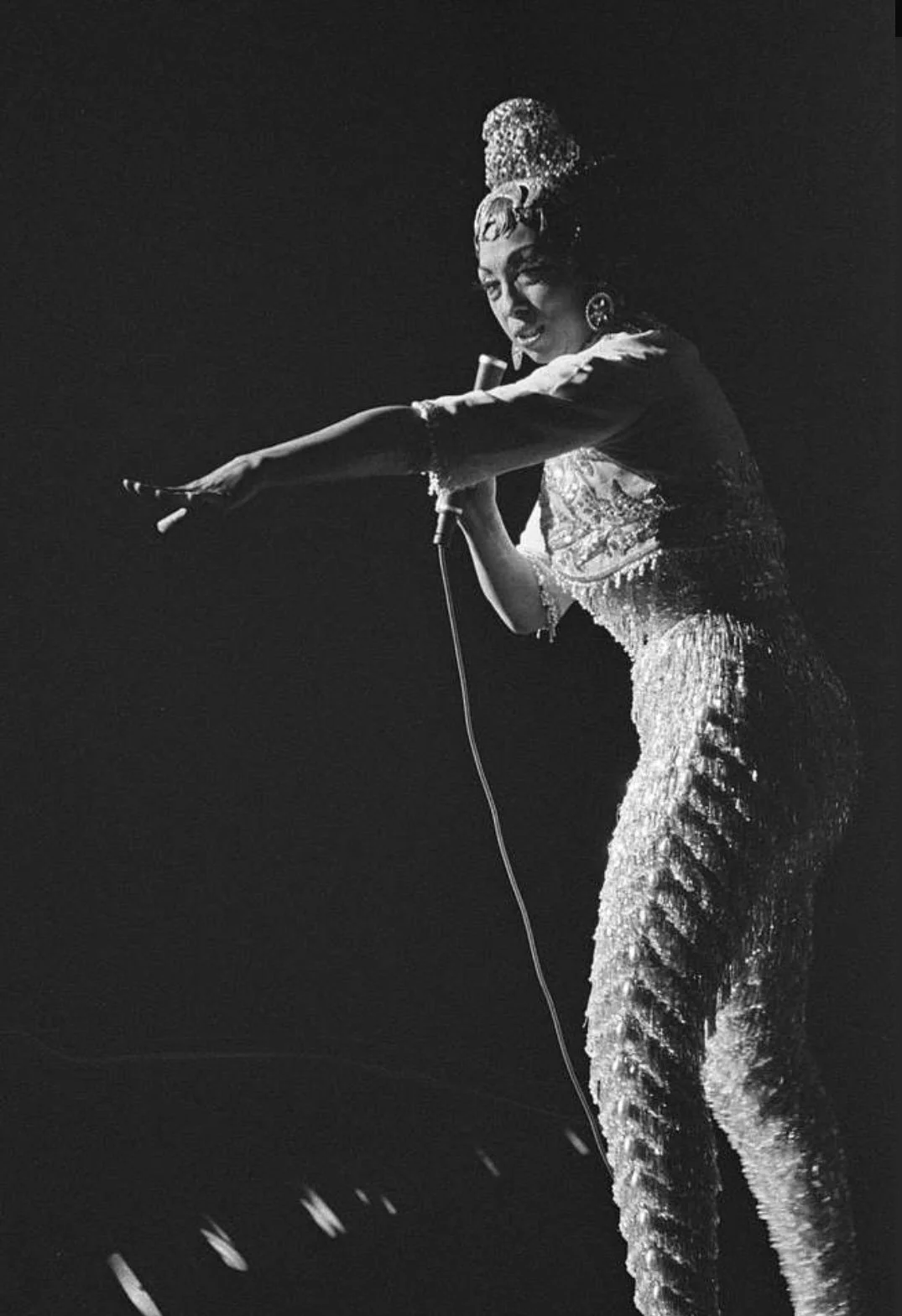 A black and white photo of Josephine Baker in elaborate costume, holding a microphone, with long fringed pants and headpiece, on stage, gesturing with one arm extended.