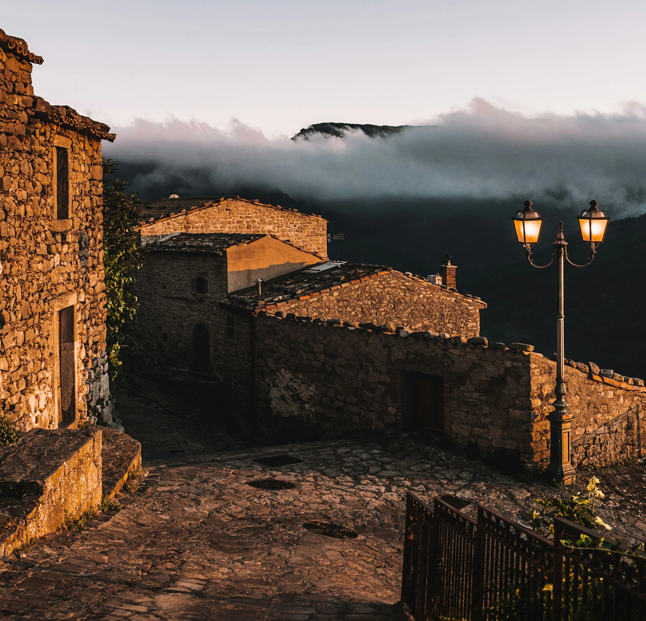A village scene at dusk with stone houses, cobblestone street, and old-fashioned street lamps, with mountains shrouded in clouds in the background.