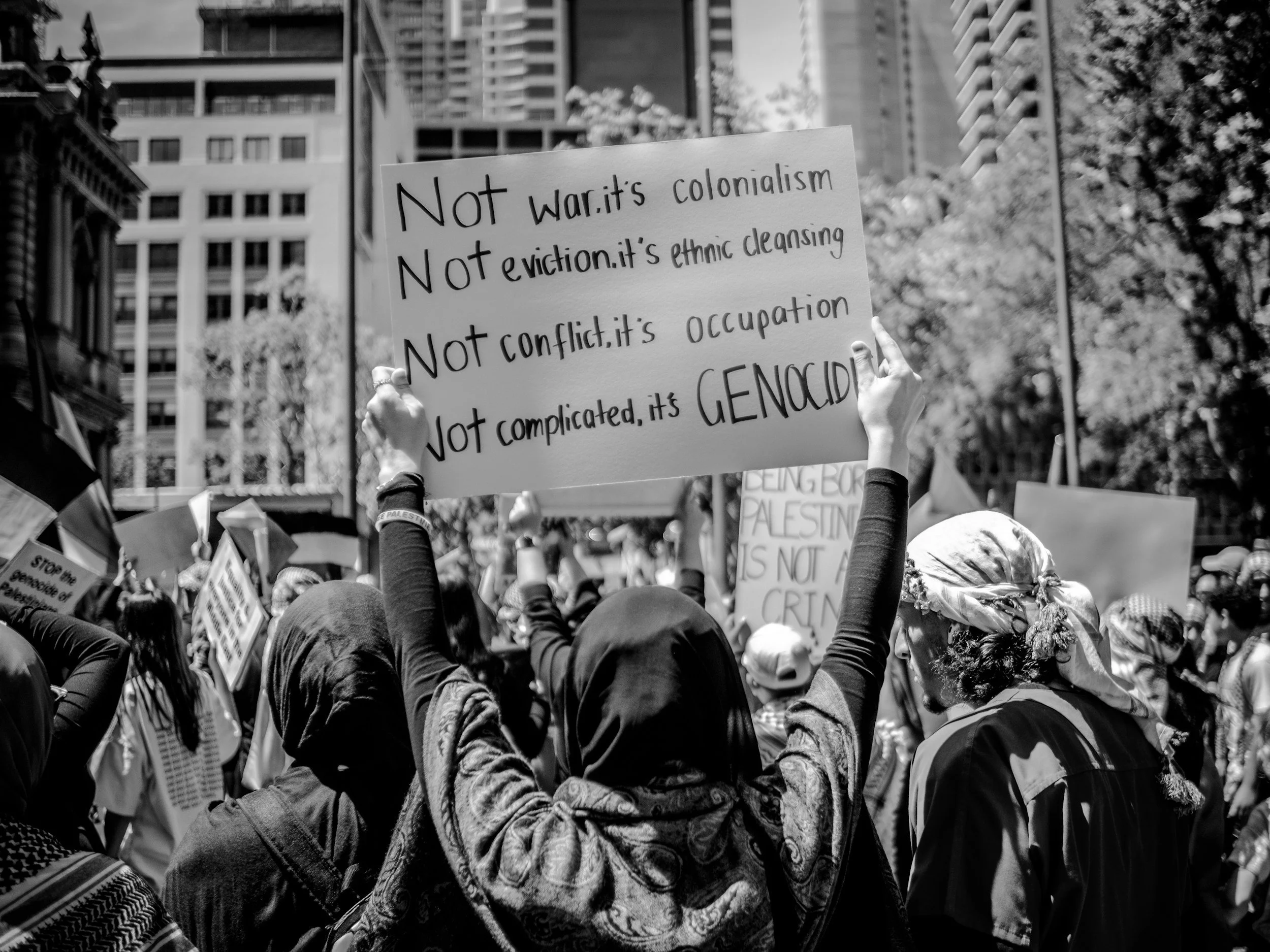 A protest march in an urban area with high-rise buildings, where a person holds a sign that reads 'Not war: it's colonialism, Not eviction: it's ethnic cleansing, Not conflict: it's occupation, Not complicated, it's genocide'.