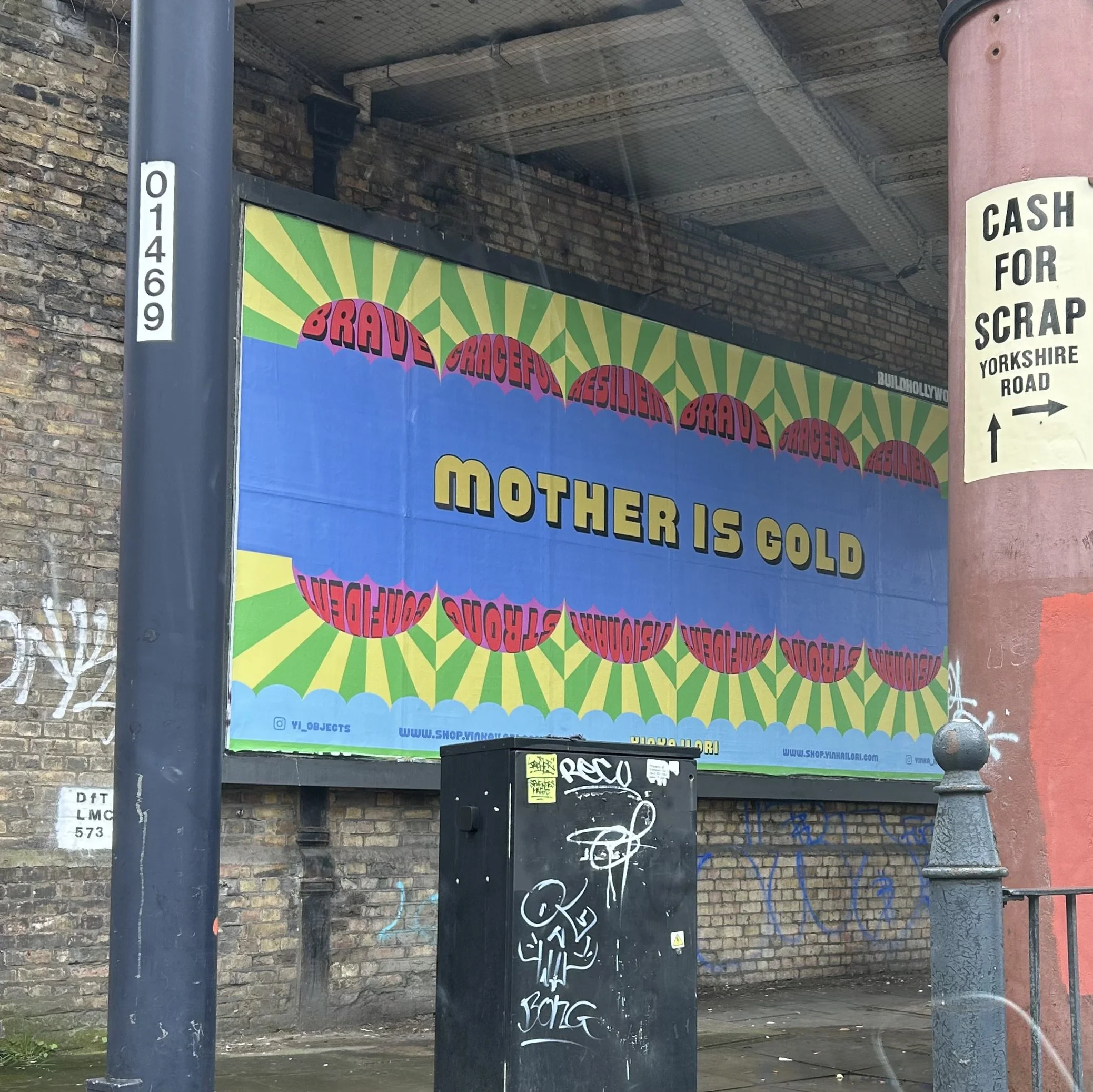 A street scene with a brick wall, and on the wall is a big advertising board that reads 'mother is gold', against an overpass or bridge with visible metal grating.