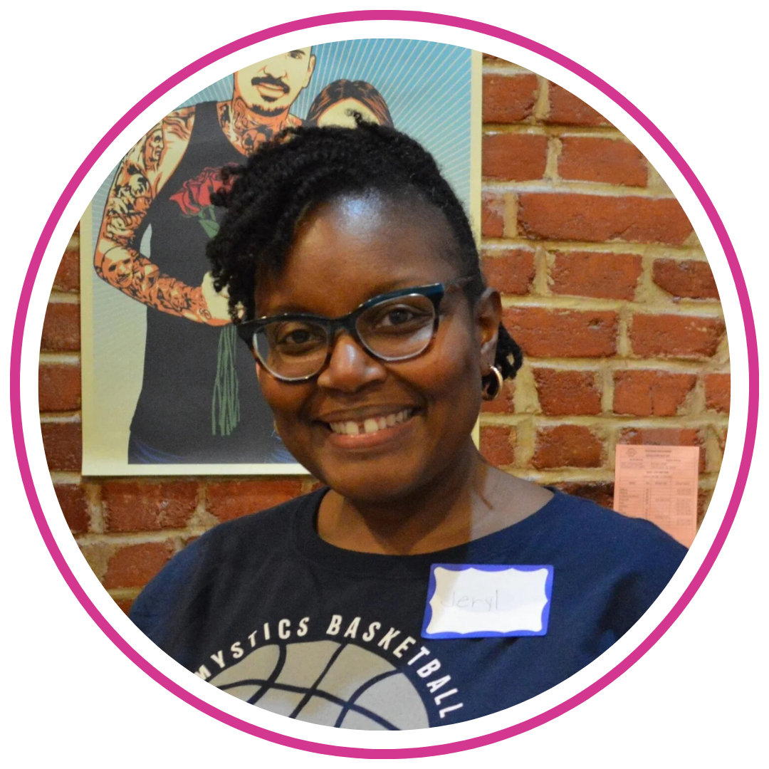 A smiling woman with short curly hair, glasses, earrings, and a navy Mystics basketball shirt, standing in front of a brick wall with a poster of a tattooed person.