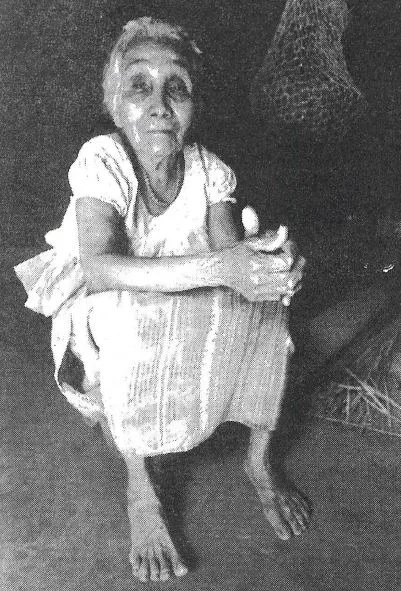 Older woman sitting on the floor, arms crossed on her knees, looking at the camera in black and white.