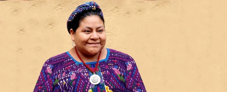 A woman wearing traditional clothing and a necklace with a medallion, smiling against a plain yellow background.