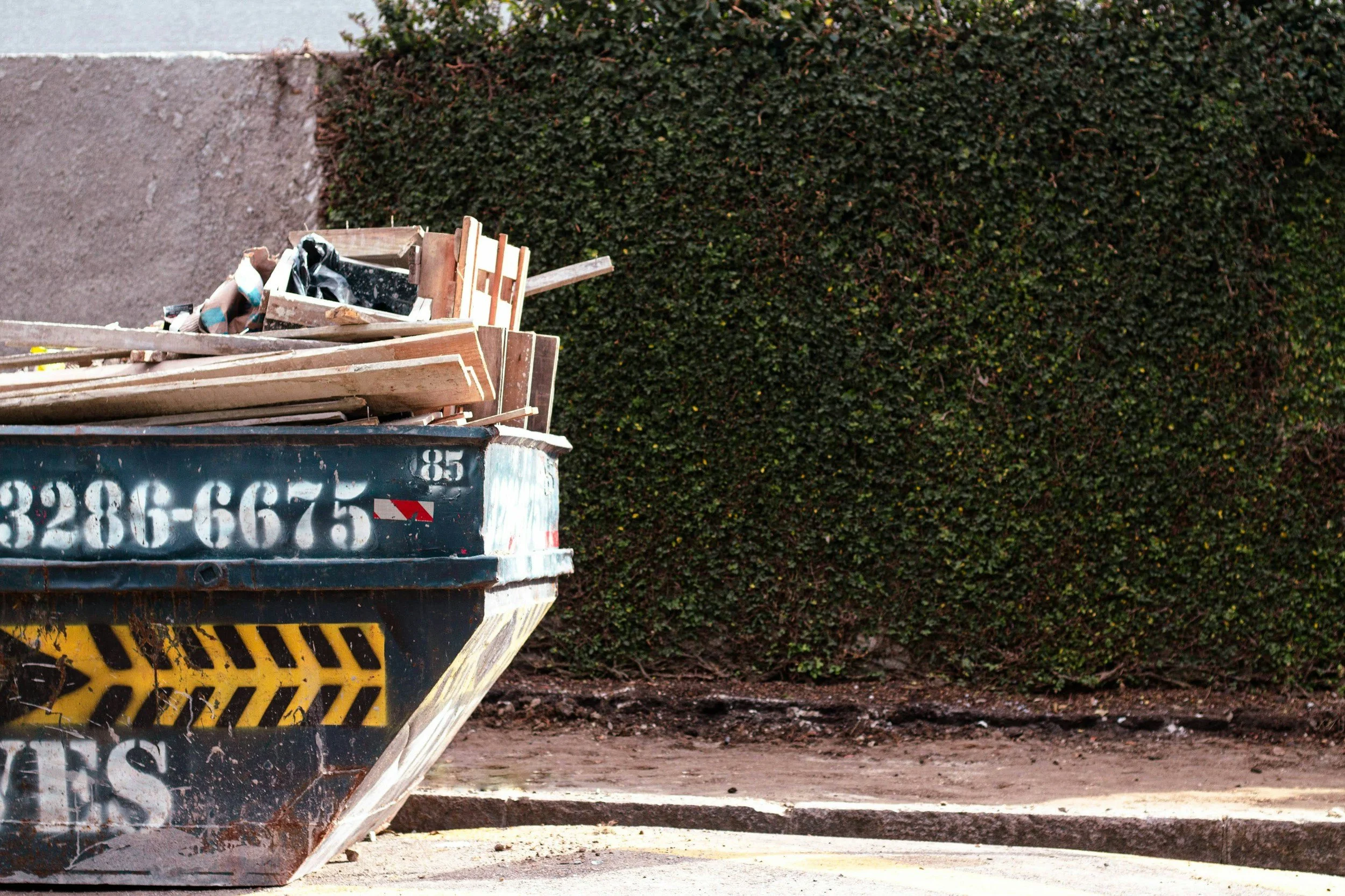 An overflowing black trash dumpster with yellow and black caution stripes on the bottom edge, filled with wooden planks, debris, and black trash bags, situated on a dirt surface next to a green hedge.
