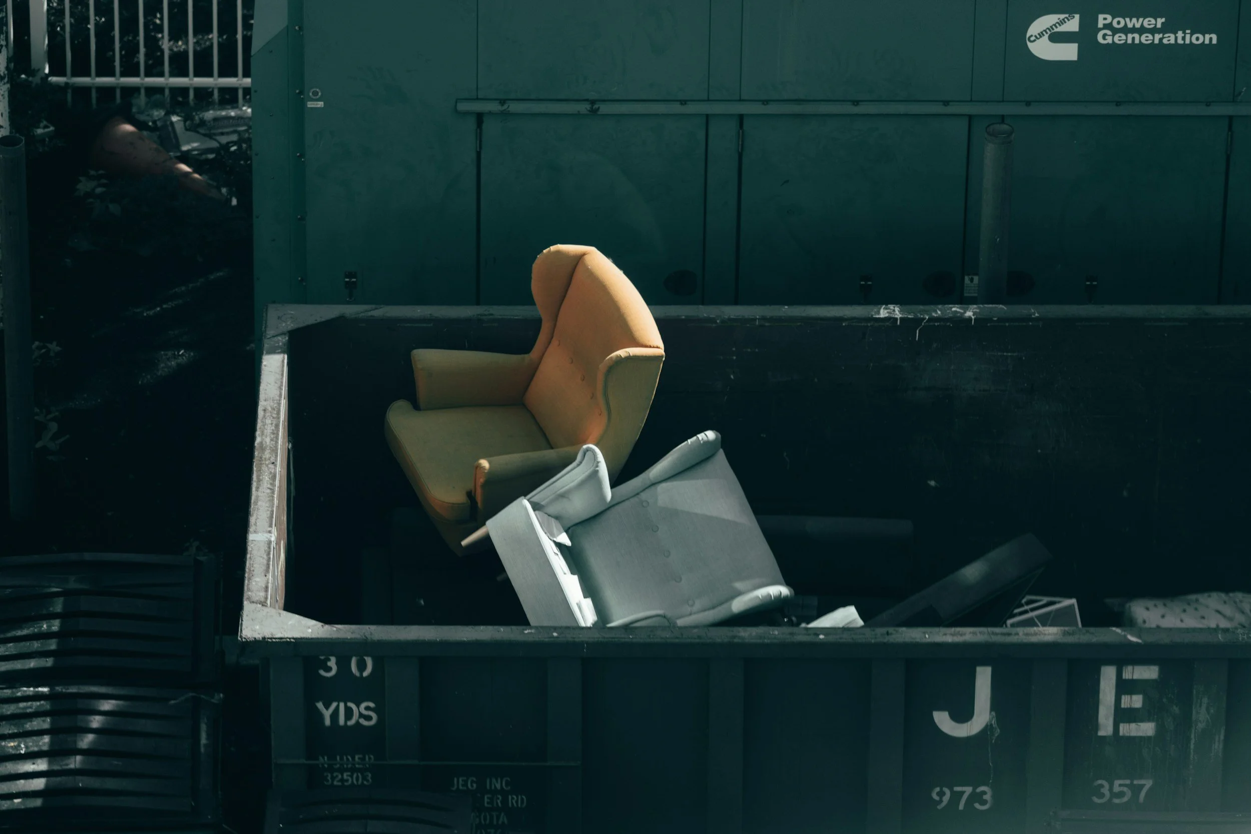 An orange armchair with a high backrest and rounded armrests sitting on a large vehicle carrier or truck bed, surrounded by other furniture and objects, with a green industrial container or structure in the background.