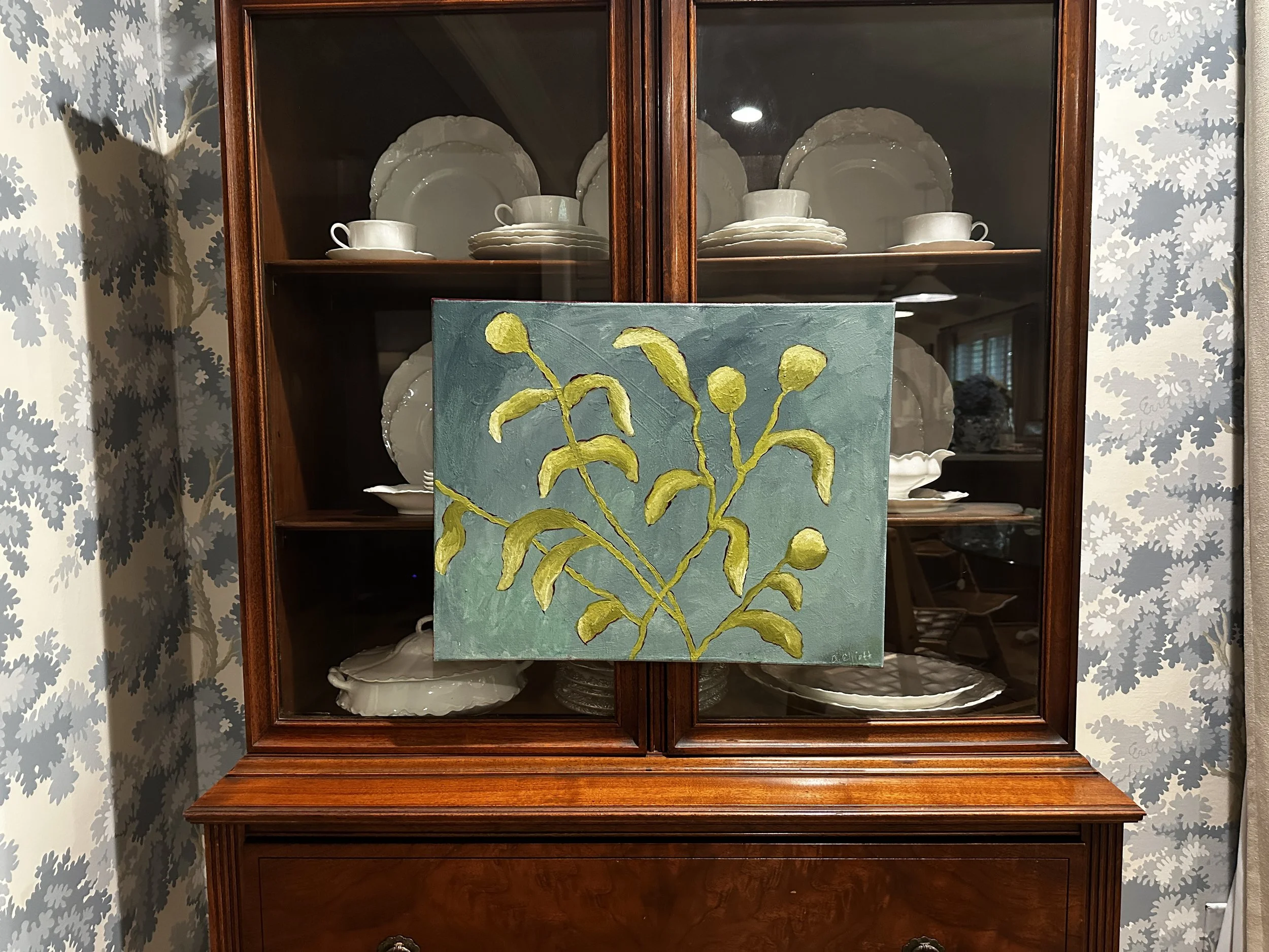 A wooden china cabinet with glass doors displaying white dinnerware and plates inside. In front of the cabinet, there is a painting of a yellow-green plant on a blue background.