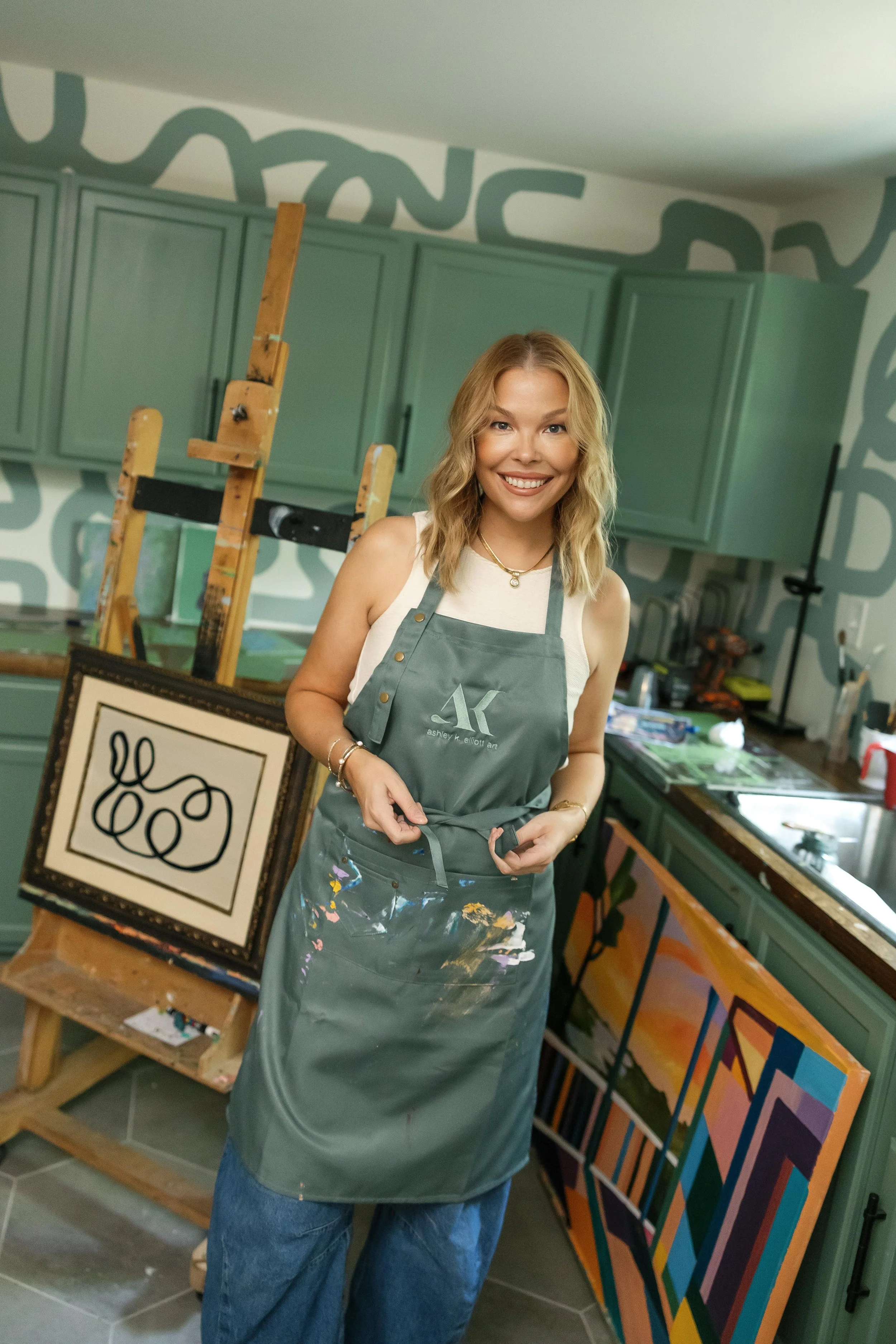 A smiling woman with blonde hair wearing a gray apron with colorful paint stains, standing in an art studio with paintings, an easel, and green cabinets in the background.
