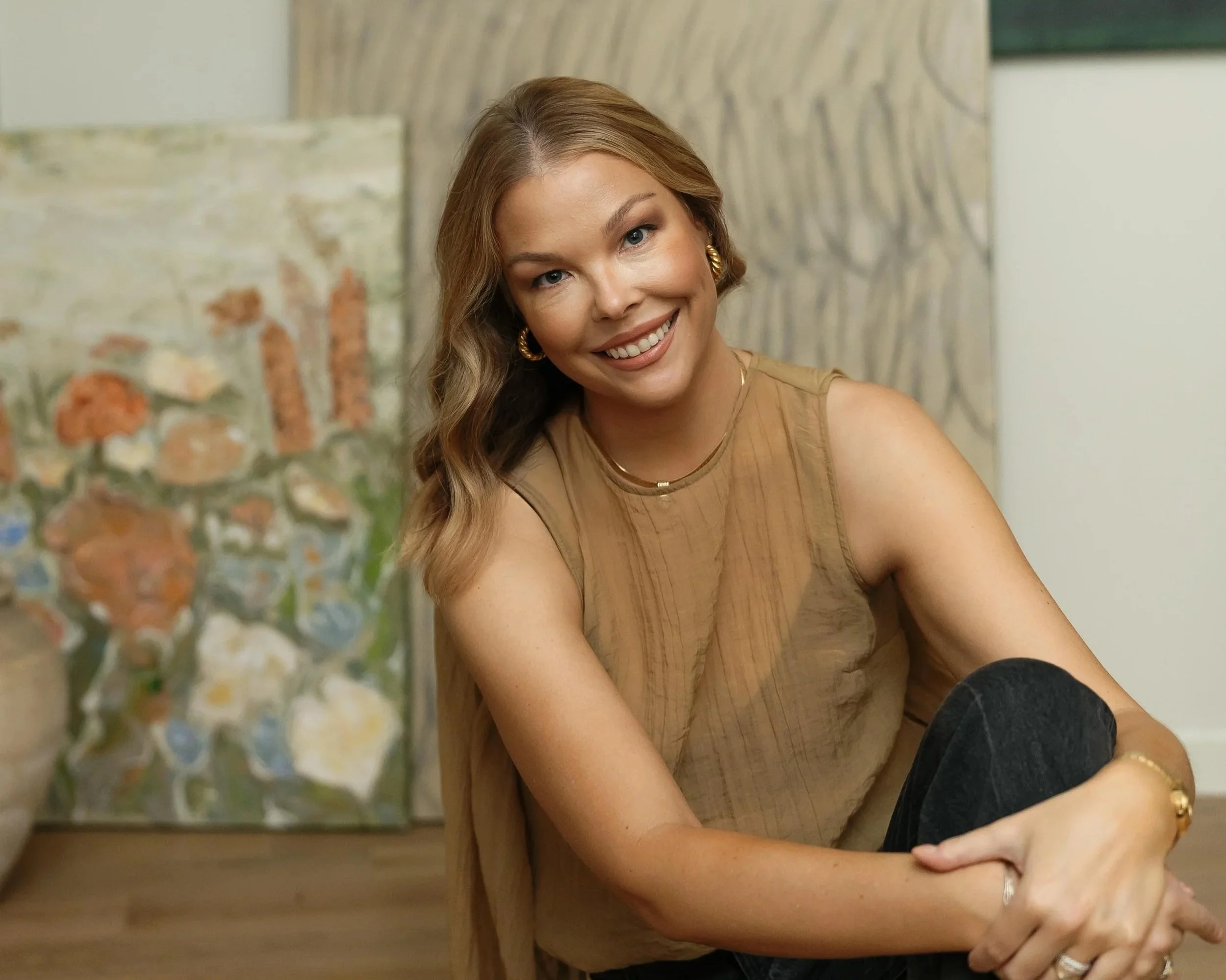 Woman with long wavy hair smiling, sitting indoors, with a decorative pillow and textured wall behind her.