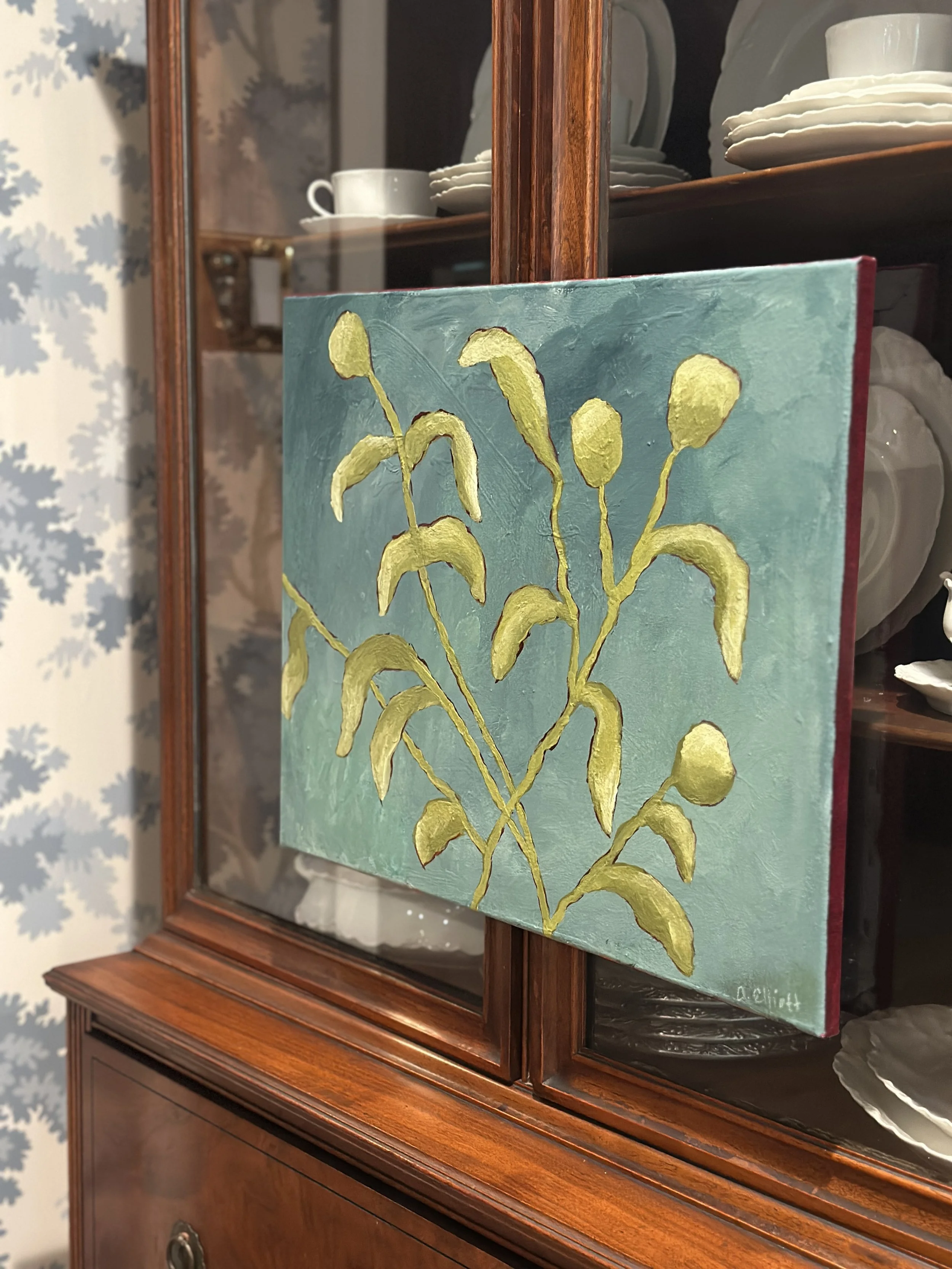 A wooden display cabinet with glass doors showcasing white china cups, plates, and bowls. A small painting of yellow-green foliage on a turquoise background is leaning against the cabinet.