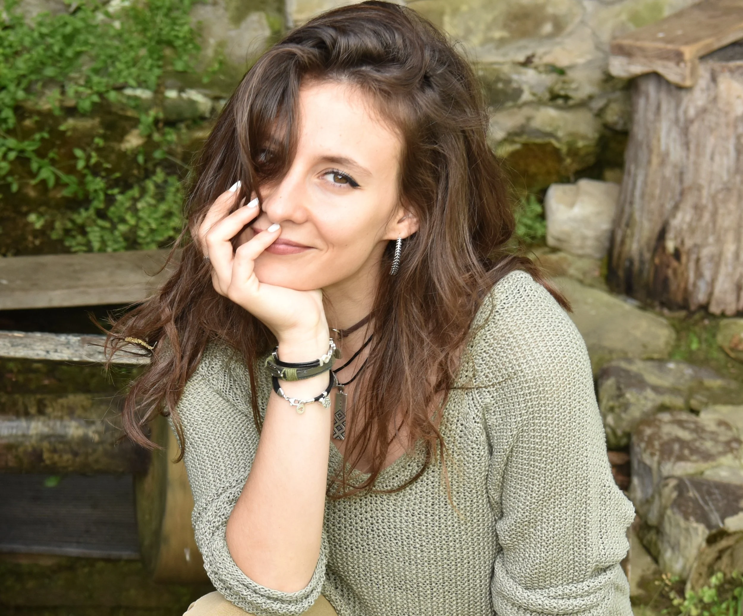 A young woman with long, wavy brown hair, wearing a beige sweater, smiling playfully near her face with her hand touching her lips, outdoors with green foliage and stone surroundings.