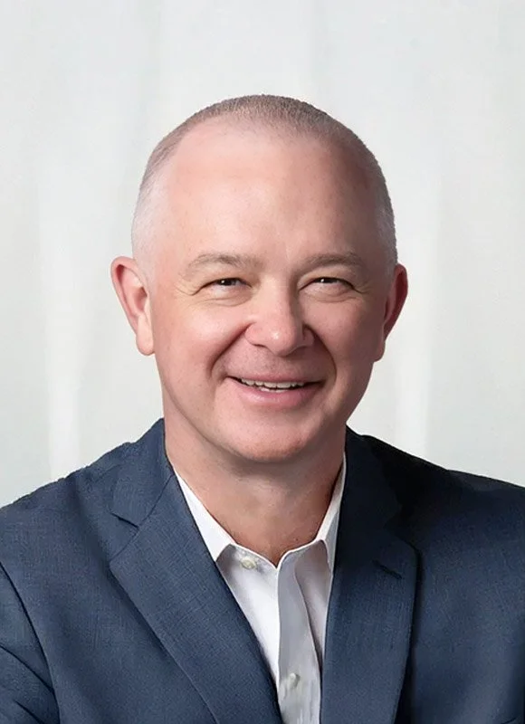 Steve Toch, a smiling man with a wearing a navy blue suit and white shirt, standing against a neutral background.