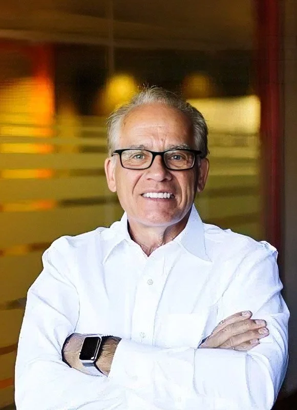 Gary Pedersen, a smiling man with glasses and gray hair, wearing a white button-up shirt, standing indoors with blurred background lighting.