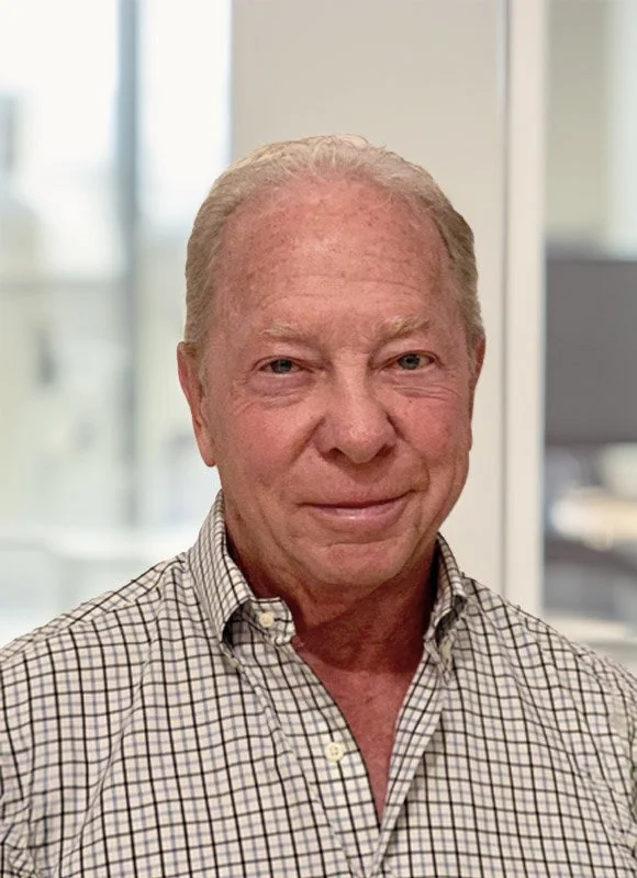 Ted Yoch, founder of We Invest In You, a smiling older man with light skin and short, light-colored hair, wearing a checkered button-up shirt, standing indoors in front of a blurred window background.