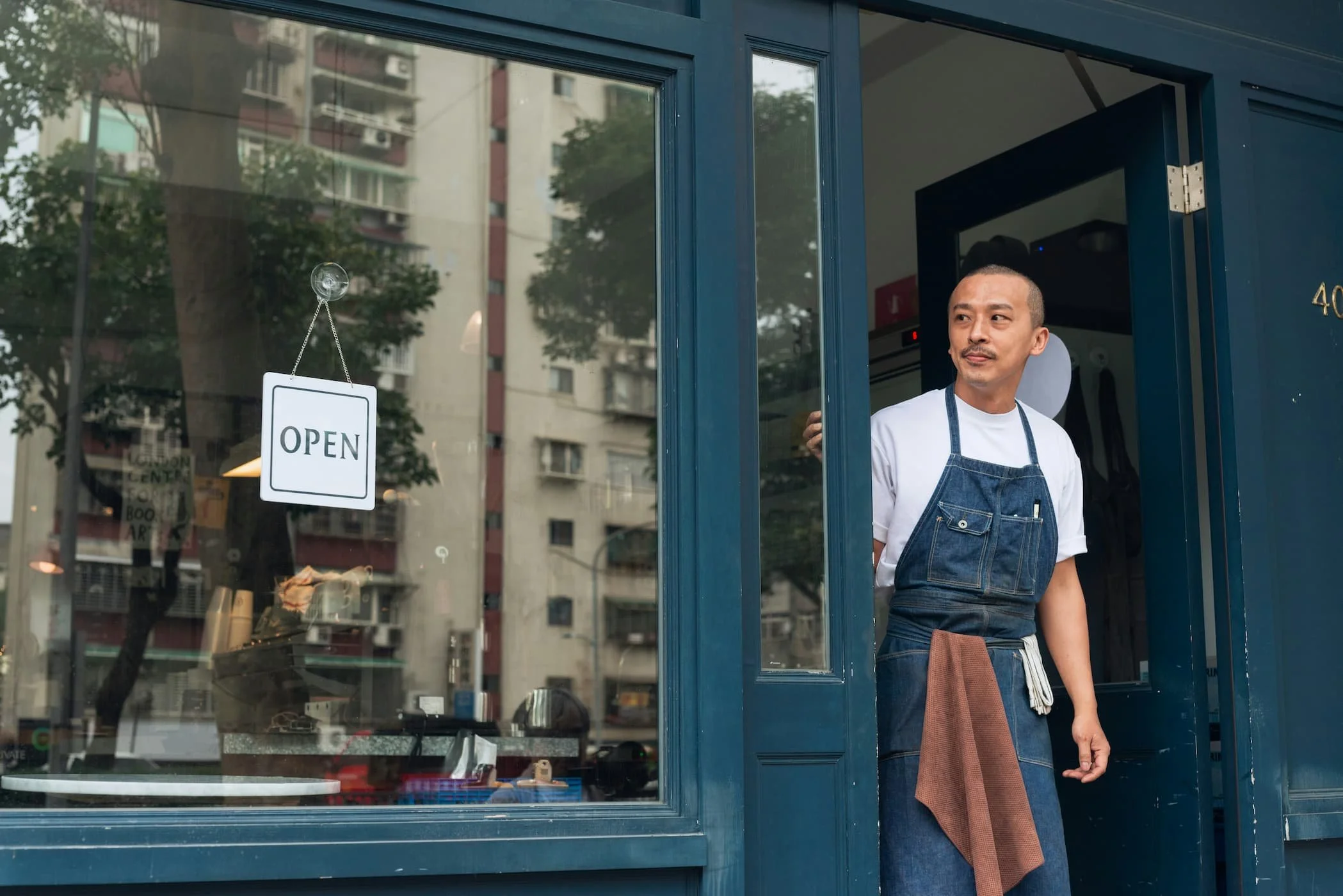 Man standing in the doorway of a cafe, wearing a white shirt and denim apron, with a brown towel over his arm, looking outside through the open door. The cafe has a glass window with an 'OPEN' sign hanging on it, and the building exterior is painted dark blue.