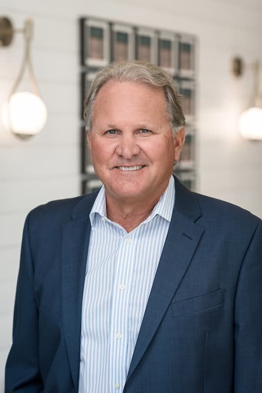 Tom Klingen, a smiling man with gray hair in a business suit, standing indoors with framed pictures on the wall behind him and wall-mounted lamps on either side.