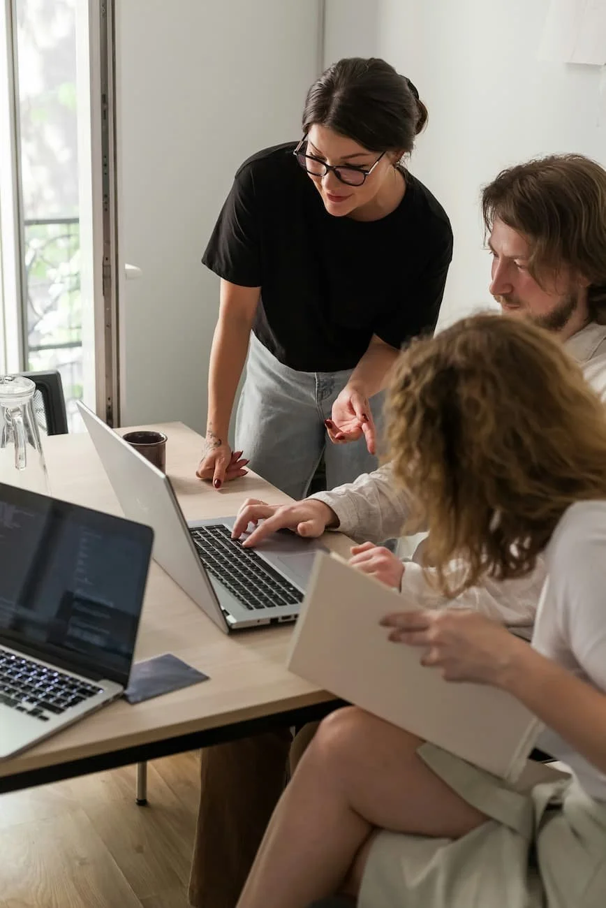 Three people gathered around laptops and a notebook, engaged in a discussion in a well-lit room with a window and a door in the background.