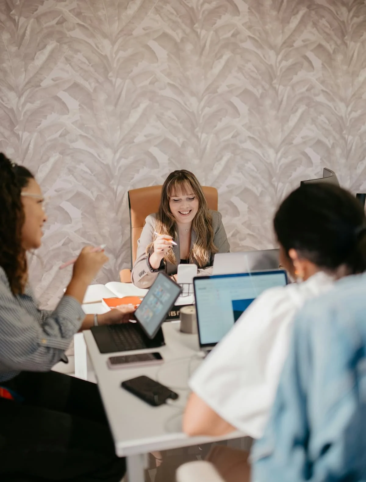 Business meeting with four diverse women around a table, using laptops and tablets, with one woman smiling and engaging in discussion in a modern office setting.