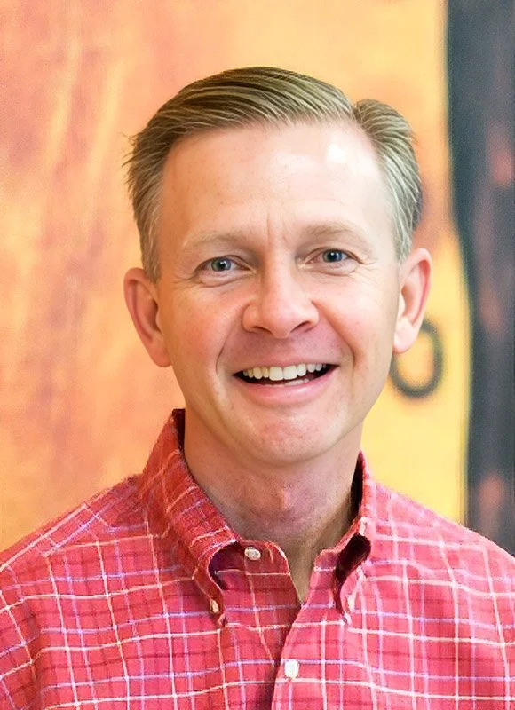 Greg Yoch, a smiling man with short, light-colored hair, wearing a red plaid button-up shirt, standing in front of a colorful blurred background.