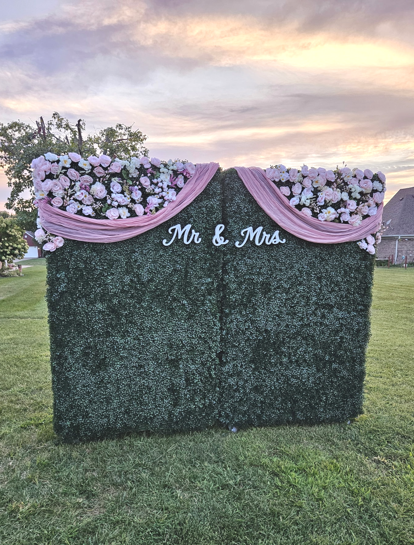 A decorated outdoor wedding backdrop with floral arrangements, pink drapery, and a sign that says 'Mr & Mrs' on a dark green hedge wall.