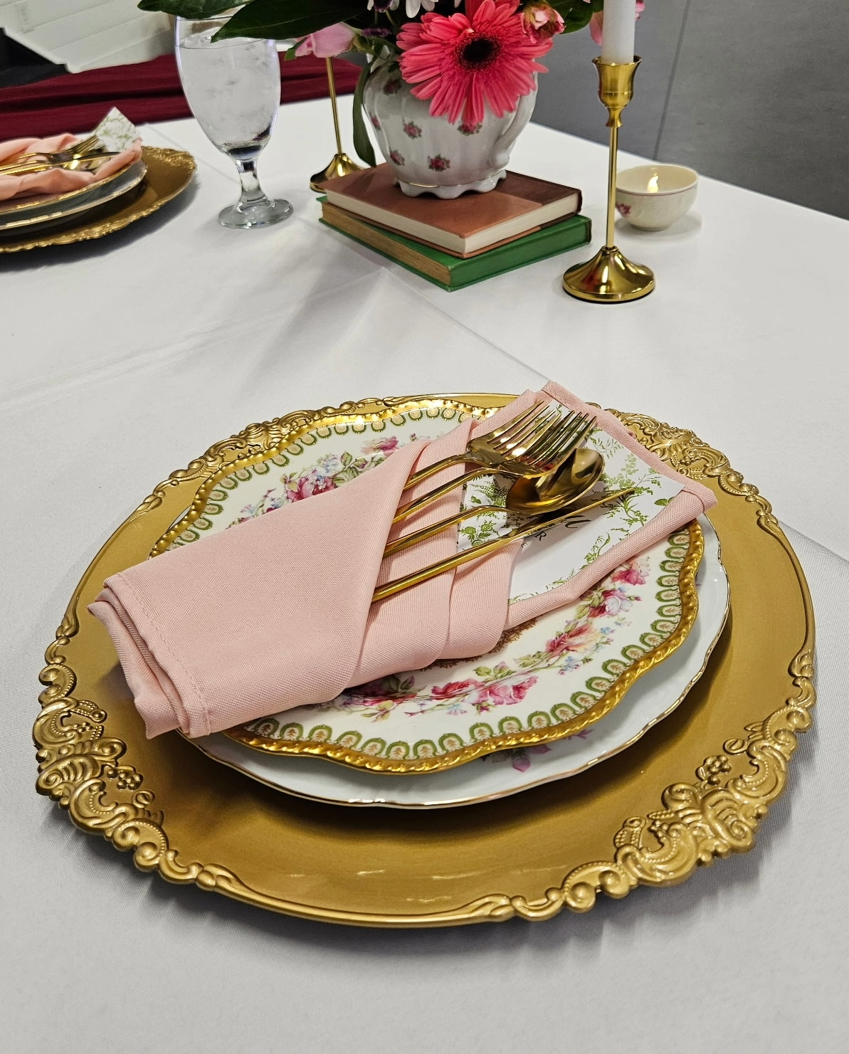 Elegant table setting with ornate gold-rimmed plates, pink napkin, gold flatware, floral decorative plate, white tablecloth, and floral centerpiece including candles and books.