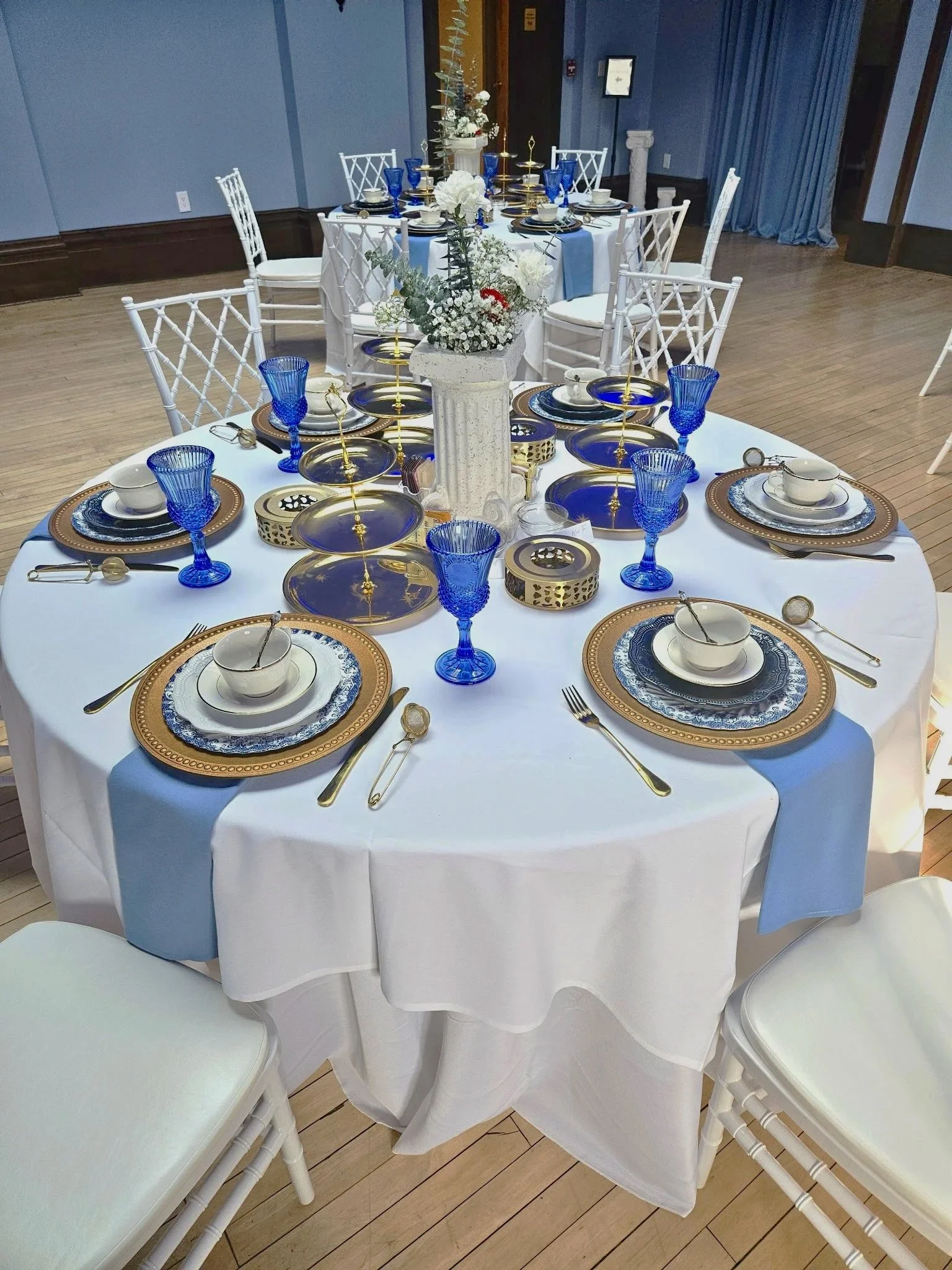 Round banquet table set for a formal event with elegant blue and gold plateware, crystal blue glasses, gold utensils, white bowls, a white tablecloth, a floral centerpiece with white flowers and greenery in a tall white vase, and a white bridal-style chair at each place setting.