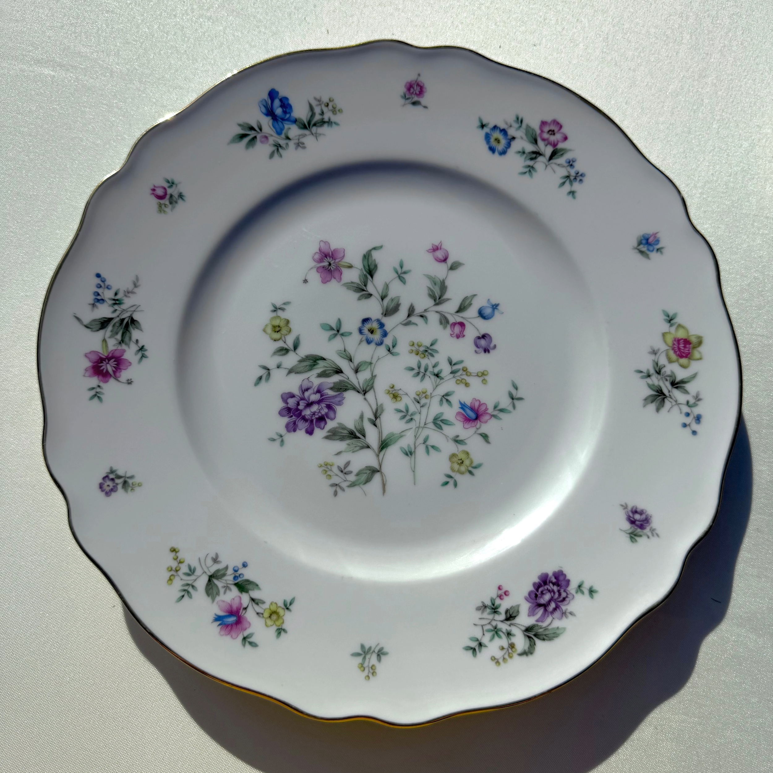 A close-up of a white, scalloped-edge porcelain plate decorated with colorful floral patterns in pink, purple, blue, and yellow with green leaves, on a white surface with sunlight casting shadows.