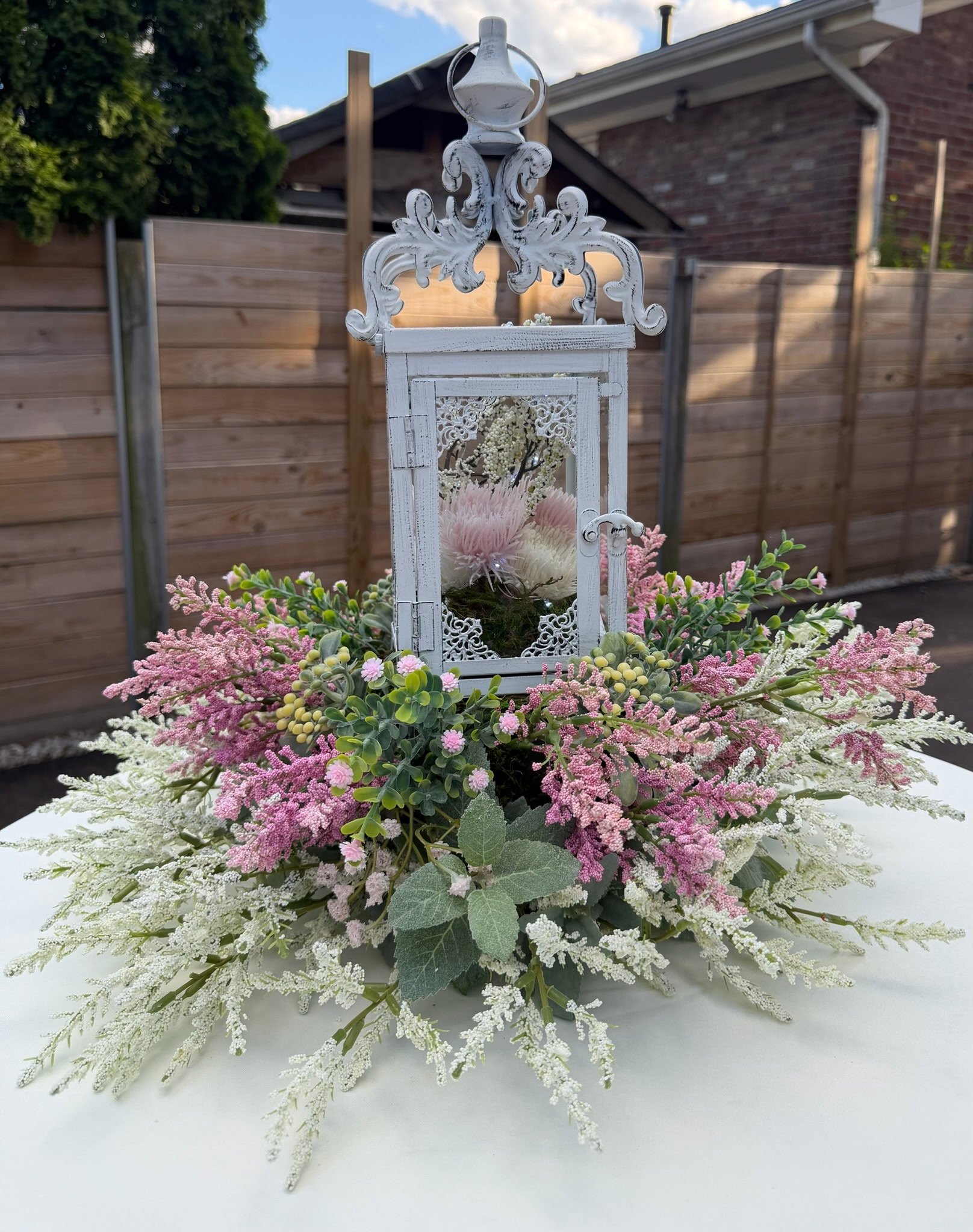 A decorative lantern filled with white and pink flowers, surrounded by additional pink and white floral arrangements on a white surface.