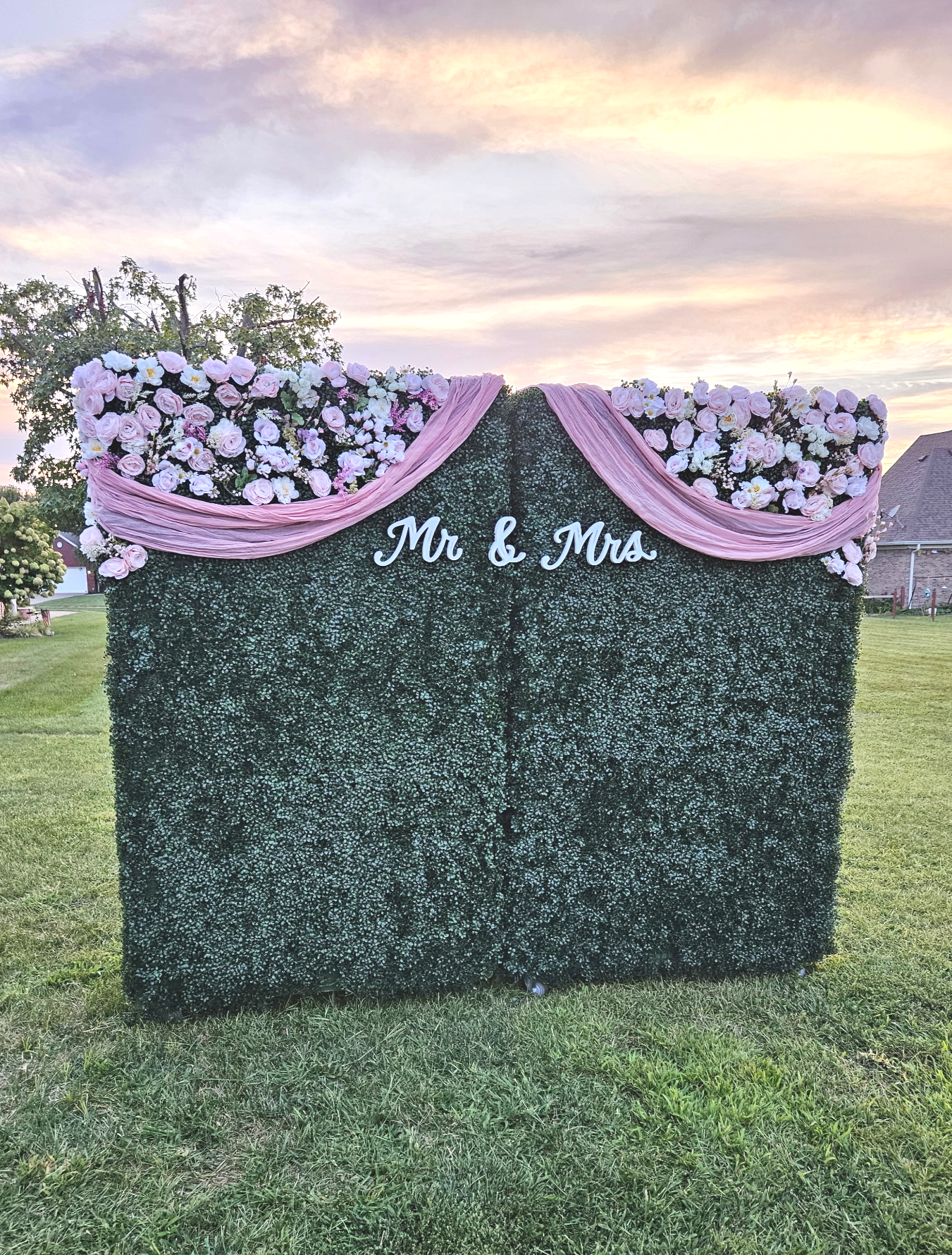 A decorative outdoor display consisting of a large artificial hedge with pink and white floral arrangements on top, draped with pink fabric. In the center, there is a sign that reads 'Mr & Mrs'. The scene is set on a grassy field during sunset.