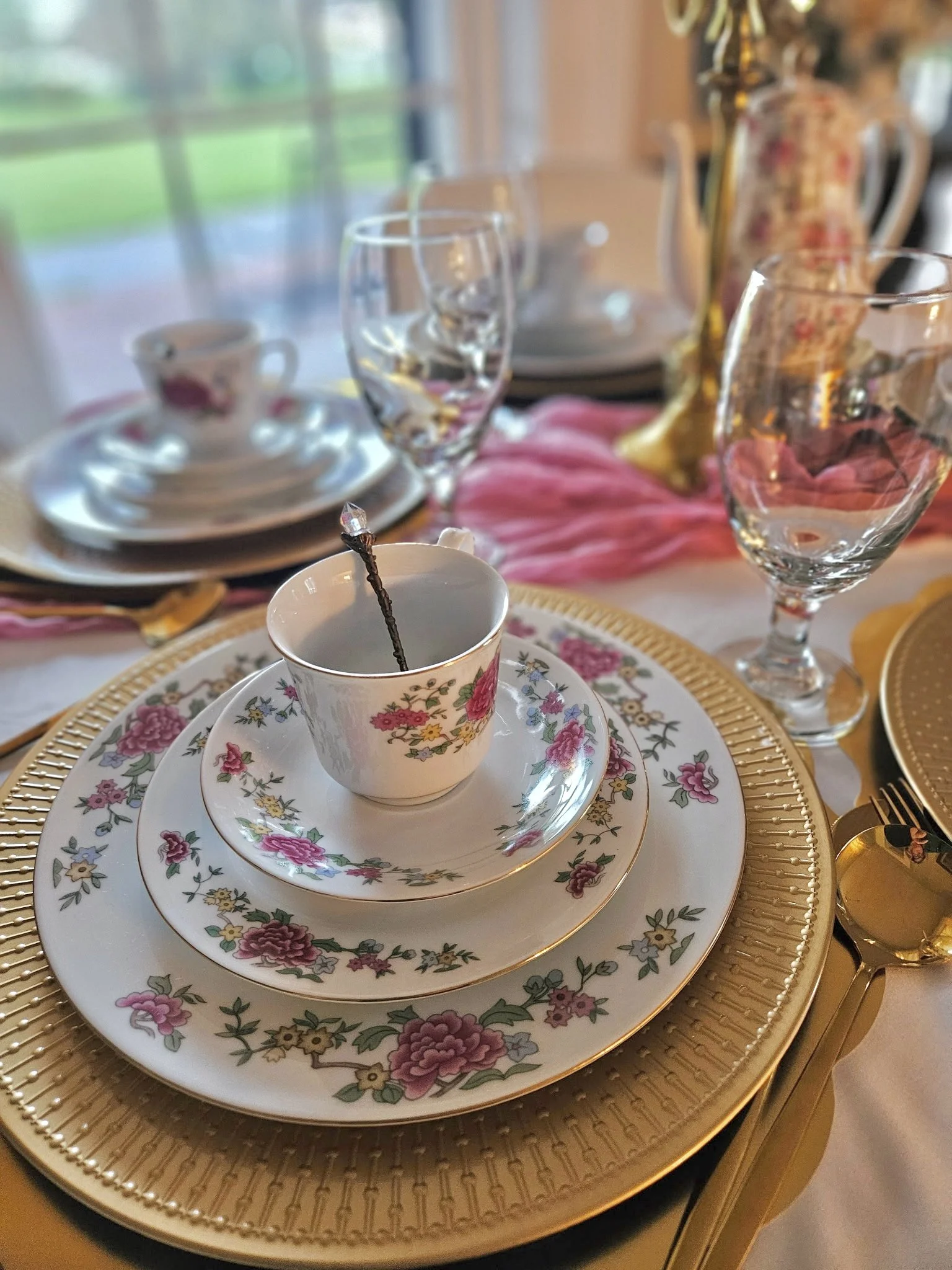 Elegant table setting with floral china teacup and saucer, gold-edged plates, crystal glasses, pink tablecloth, and gold accents, near a window with a blurred outdoor view.