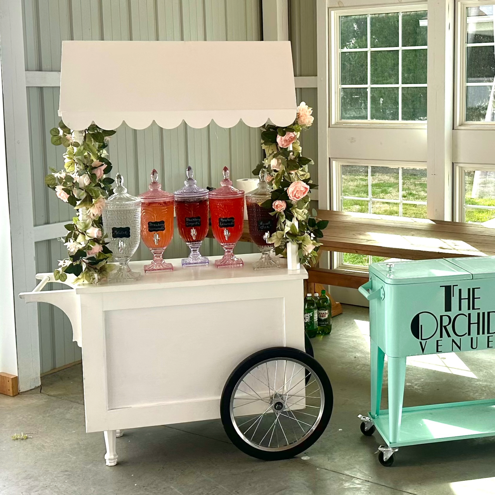 A white beverage cart with a decorative canopy, adorned with pink and green flowers on the sides, displaying four large glass dispensers with beverages, standing inside a bright room with large windows.