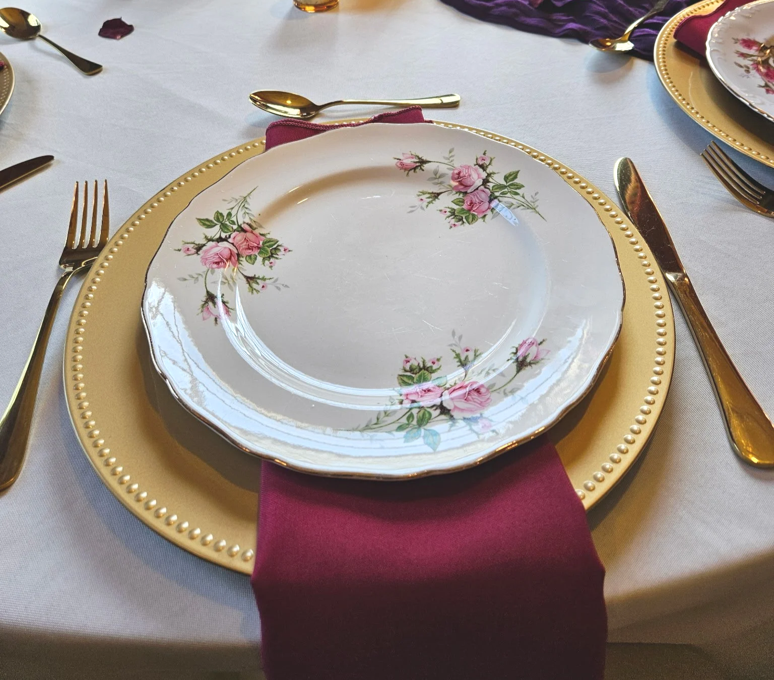 A table setting with floral china, gold flatware, and a burgundy napkin.